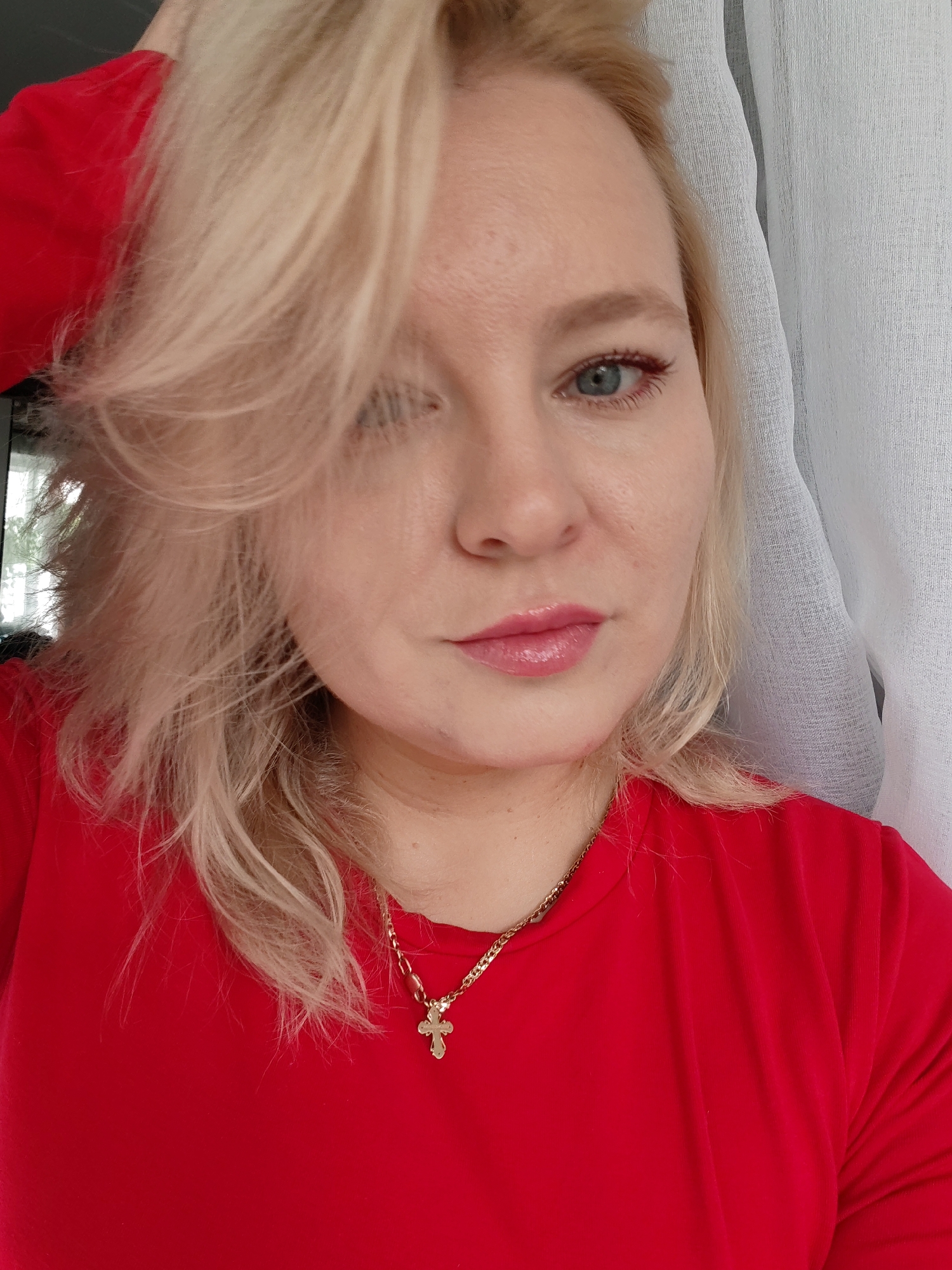 A close-up, shoulder-up shot shows a blonde woman with blue eyes and light pink lipstick. She is wearing a bright red shirt and a gold chain with a cross pendant. Her hair is styled casually, with strands falling around her face. A sheer white curtain is visible behind her. The lighting appears to be natural, coming from the side. The image was captured in Izhevsk, Russia.