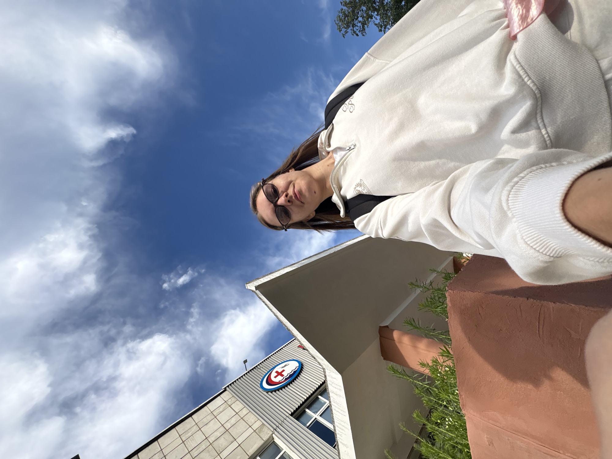 An upward-angle, wide-shot selfie shows a young woman with long brown hair, wearing sunglasses and a cream-colored quarter-zip sweatshirt with a backpack over her shoulders. She is standing outside a modern building with a unique angular roofline, in the city of Chaikovsky, Russia. The building has a distinctive circular logo with a red cross in the center on its facade. The sky above is a vibrant blue with scattered white clouds. Green foliage and a reddish-brown concrete planter are visible in the foreground.