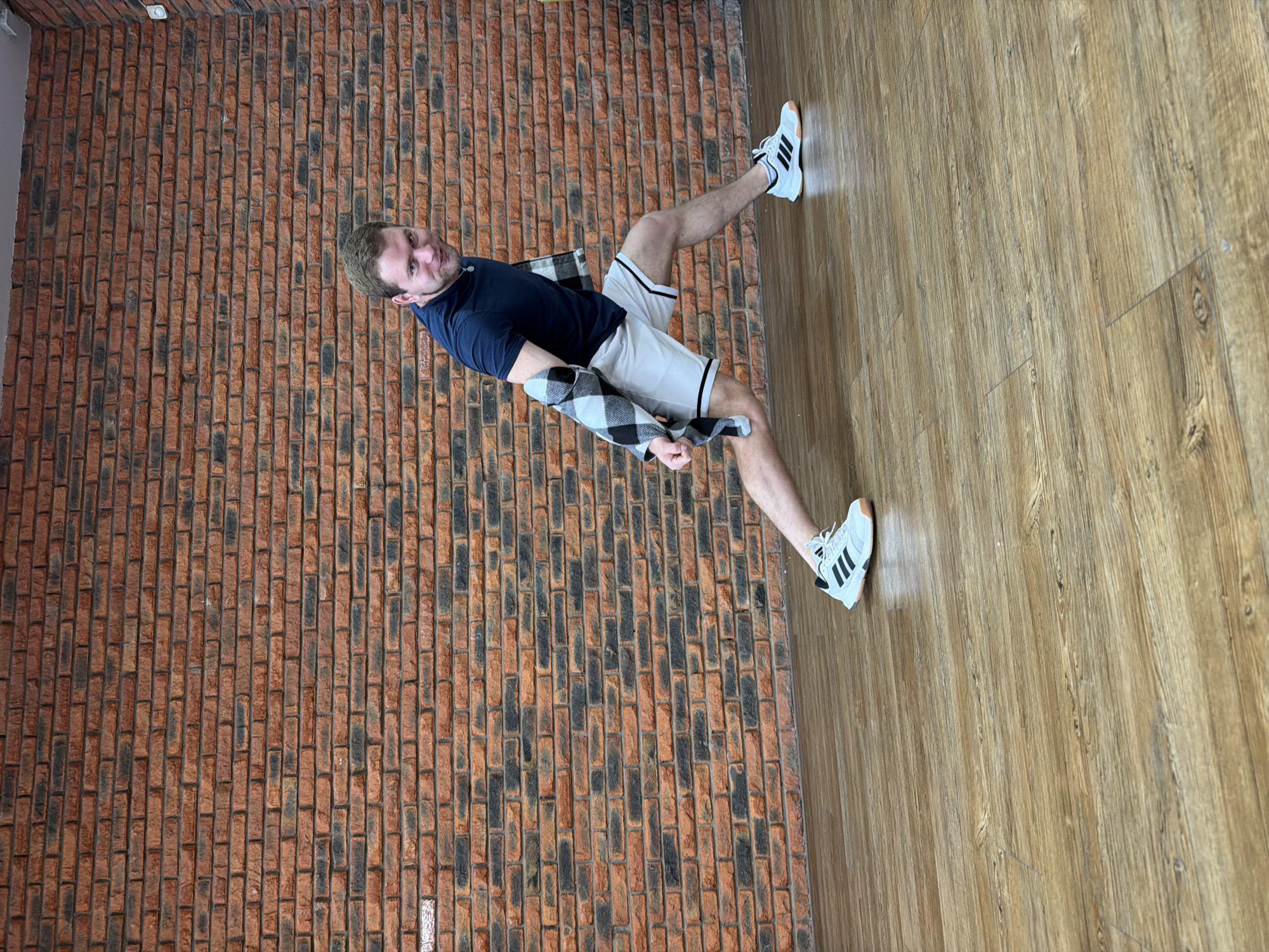 A man wearing a navy blue t-shirt, white shorts with black stripes, and white Adidas sneakers is leaning against a brick wall with a black and white checkered flannel shirt around his shoulders. The man is in an indoor setting with a wooden floor. The exact location in Chaikovsky, Russia, is not discernible from the image.