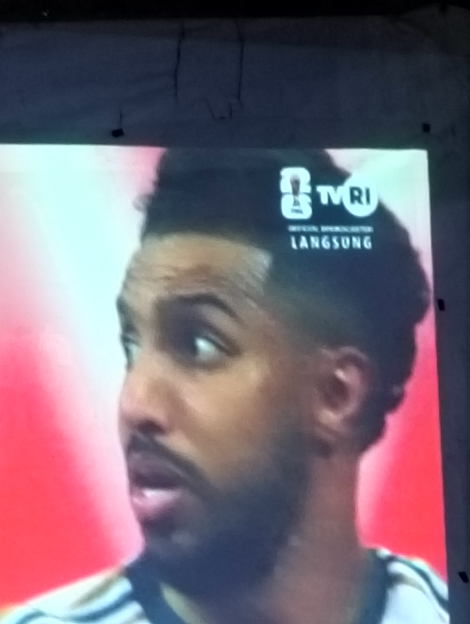 The image shows a close-up of a man's face. He has dark hair styled with a high fade. He is looking to the right of the frame. Above his head, there is a logo that reads "TVRI" with the word "LANGSUNG" underneath, which means "live" in Indonesian. The background is a blurry red and white. The man is wearing a white shirt with black stripes. The scene appears to be from a television broadcast. There is no visible indication of the city of Payagaboh, Indonesia.