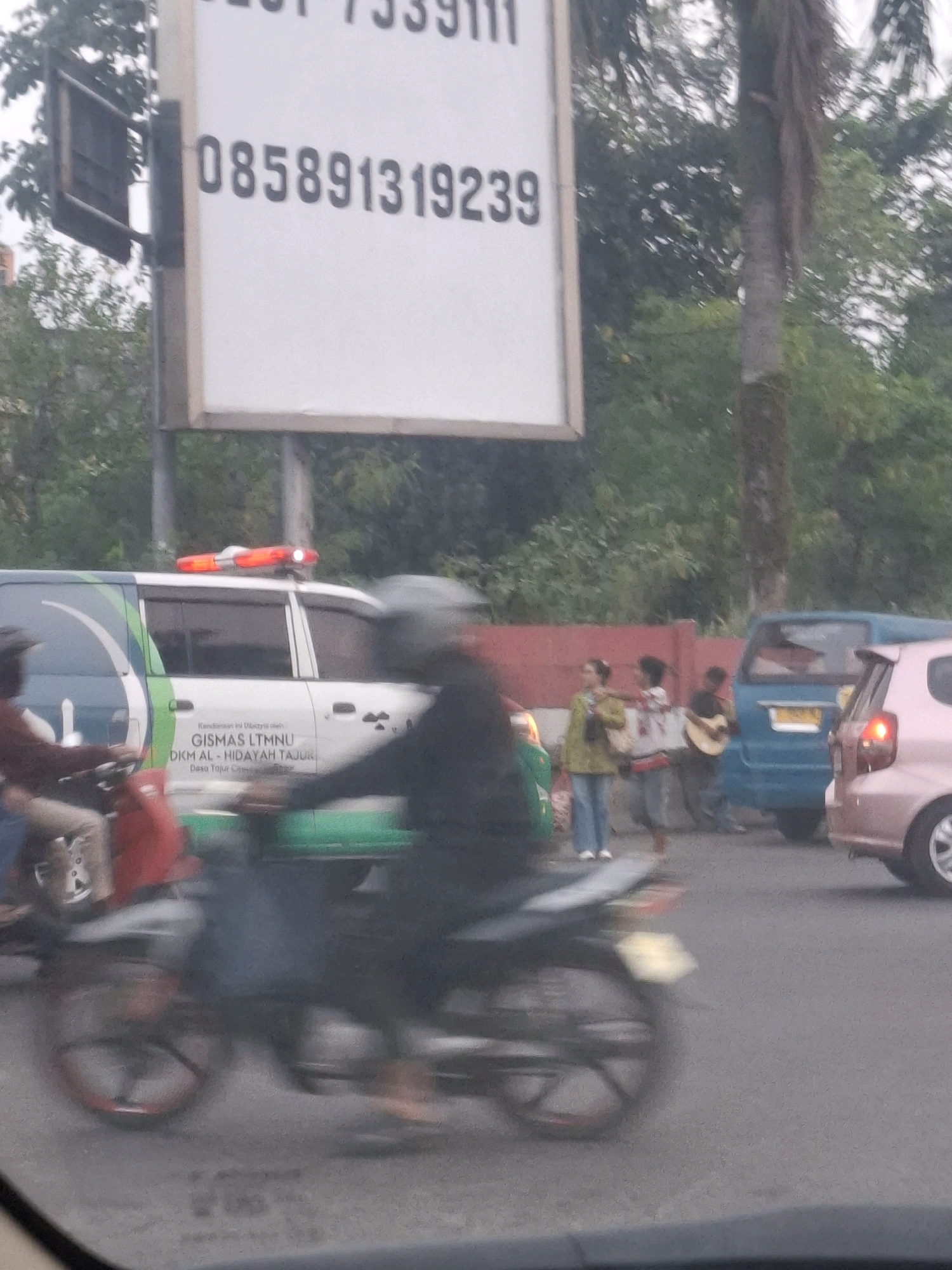 The image captures a street scene, likely in Citeureup, Indonesia, focusing on a white van with red flashing lights on its roof. The van displays text including "GISMAS LTMNU" and "DKM AL - HIDAYAH TAJUR". In the foreground, a person is riding a motorcycle, blurred due to motion. Behind the van, a blue minivan and a pink car are visible. Further in the background, a group of individuals are gathered; one person plays a guitar, another holds a bottle, and a third gestures with their hand. Trees and foliage line the street, suggesting an outdoor urban environment. A large white sign with black text is prominently positioned on the left side of the image. The overall impression is a busy street with various vehicles and pedestrians.