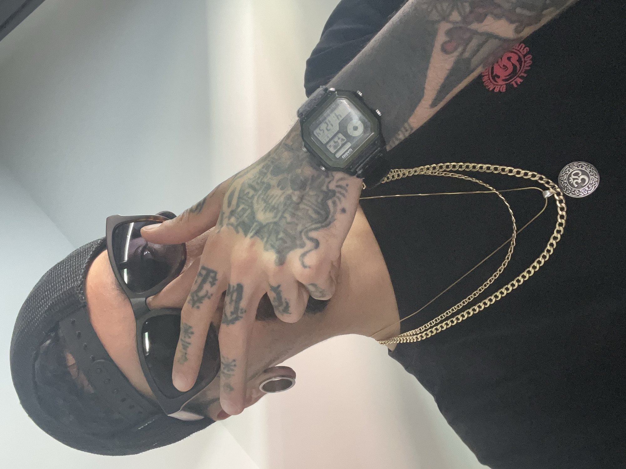 A man is taking a selfie in a mirror. He is wearing a black t-shirt, a black baseball cap, and sunglasses. He has tattoos on his hands and arm, and he is wearing a watch and two gold necklaces. He also has an ear gauge. The background appears to be a plain wall. There is no indication of his location in Madrid, Spain.