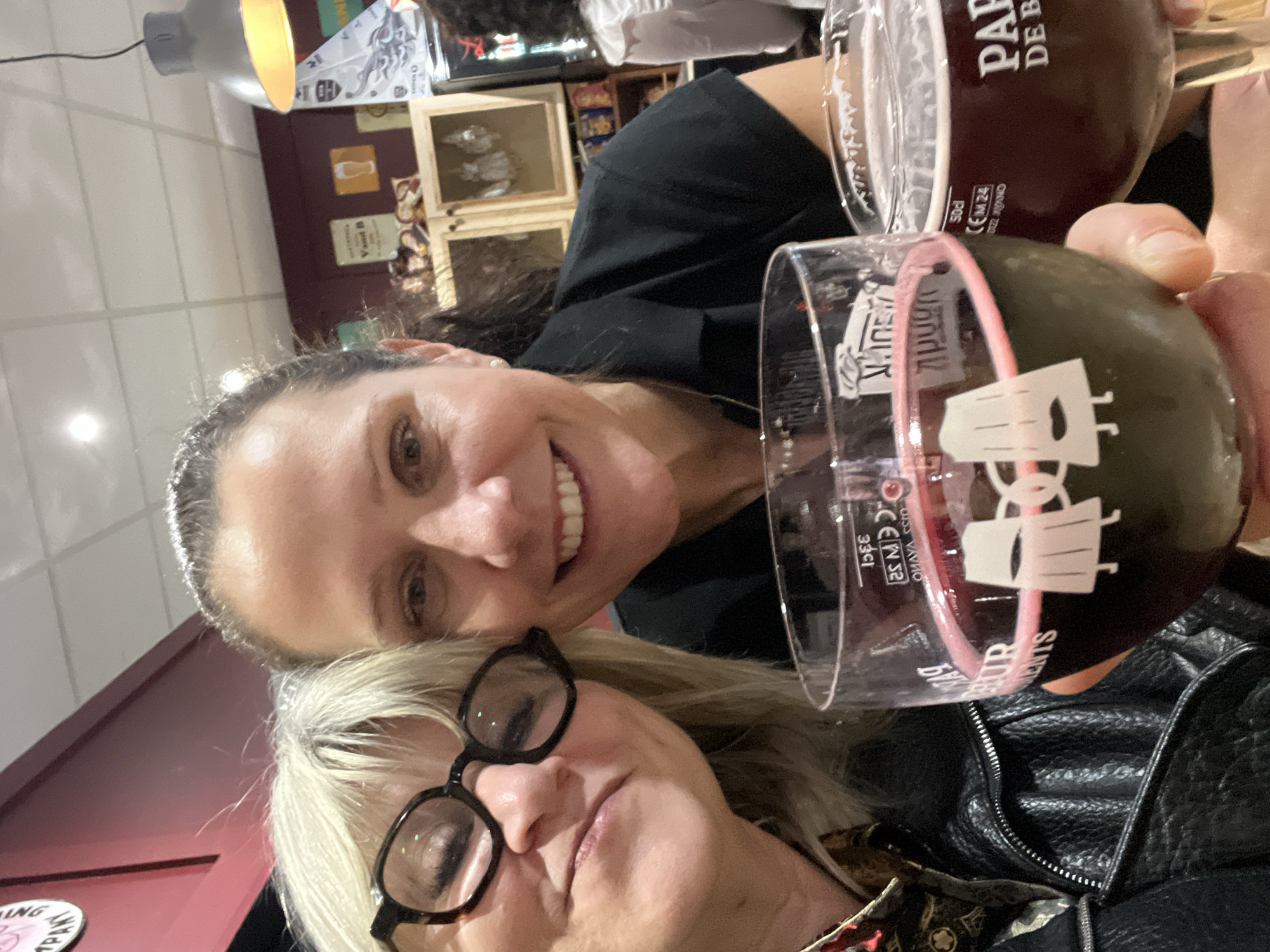 An eye-level, close-up photograph captures two adult females posing for a selfie inside a V&B (Vins & Bières) bar or pub, located in Montbrison, France.
The subject on the left, a female with blonde hair and dark-framed glasses, wears a black leather jacket over a patterned shirt. Her left eye is partially closed, and she has a faint smile. The subject on the right, a female with dark hair pulled back, wears a black collared shirt and displays a broad smile. Both individuals are looking directly at the camera.
Each woman holds a large, stemmed glass filled with a dark reddish-brown liquid, consistent with beer, exhibiting a foamy head. The glass held by the left subject features a white graphic of two stylized beer steins "cheers-ing" and "33cl" text. The glass held by the right subject displays "50cl" and partial branding "PAP DE BE." Their hands are positioned as if making a toast.
The background shows a dark red wall decorated with various signs, including one with "Vand B" and a beer glass icon. Wooden shelving units containing products and a dark beverage cooler are visible behind the subjects. Overhead, a grey industrial-style pendant light emits a warm light, complementing additional ceiling spotlights set into a white tiled ceiling. A third individual is partially visible in the far-right background, seated with their back to the camera, wearing a white garment.
