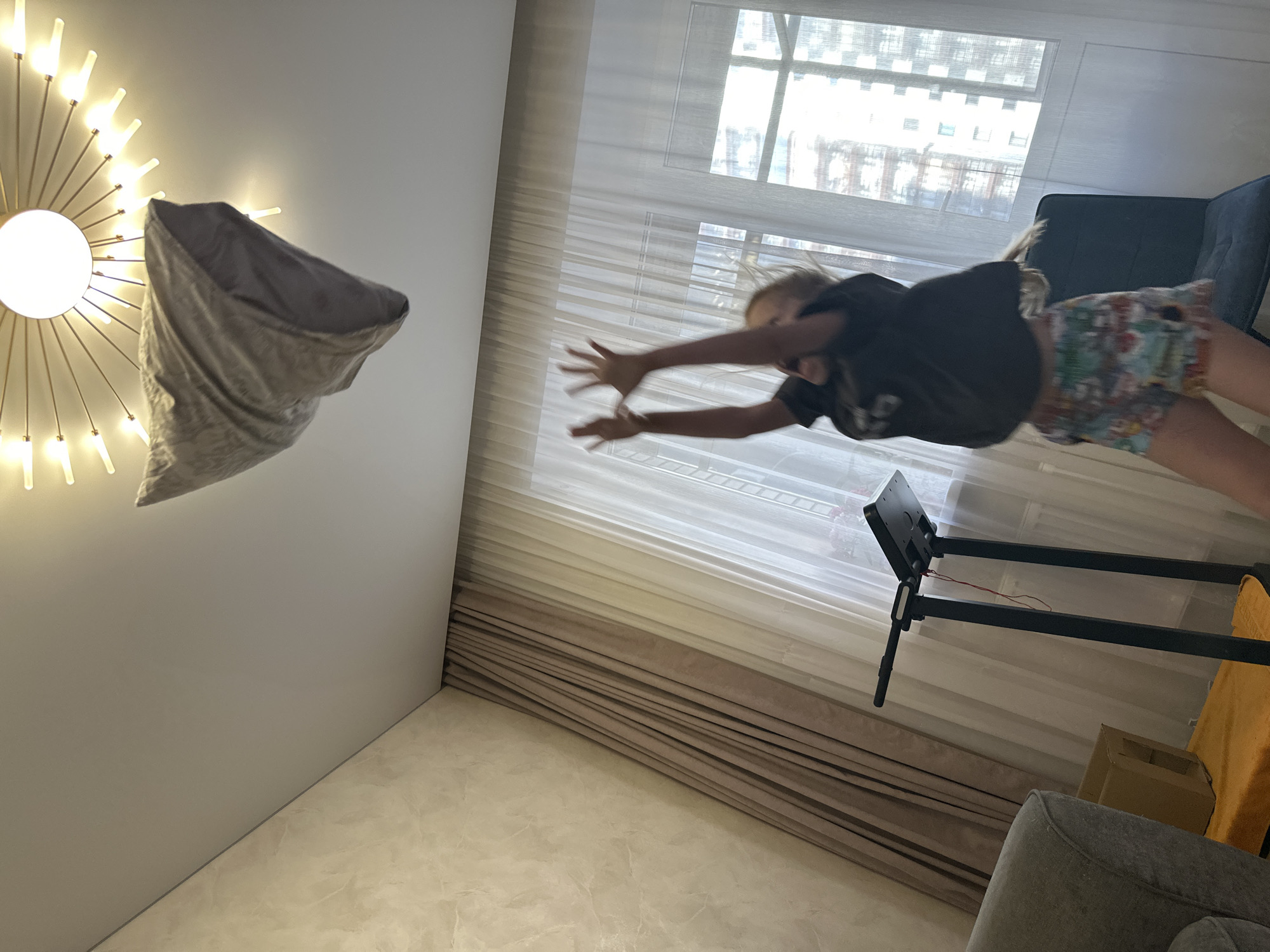 The image depicts a young child with blonde hair, wearing a dark t-shirt and colorful shorts, in mid-air, reaching upwards with both hands. The child appears to be jumping, possibly attempting to catch a pillow that is suspended in the air above them, near a modern, starburst-shaped ceiling light fixture. The setting is an indoor room with textured beige walls and floor-to-ceiling curtains. To the left, a portion of a gray sofa is visible, and further back, a treadmill with a black frame and a red cord is present. A cardboard box rests on an orange surface behind the treadmill. Through a large window in the background, the facades of several buildings can be seen, suggesting an urban environment. The lighting suggests daylight entering through the window and ambient room lighting from the fixture. No specific geographical markers are visible to confirm a location in Abakan, Russia.