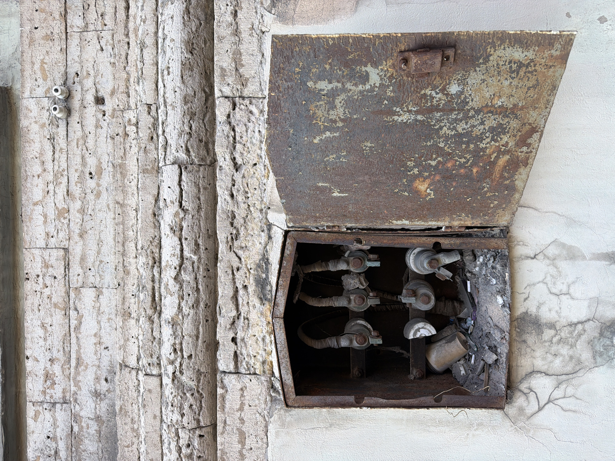 The image shows a weathered, rusty electrical box mounted on a stone wall. The box is open, revealing old electrical components and debris. Two small white ceramic insulators are visible on the wall above the electrical box. The wall itself is constructed from rough-hewn stone blocks, showing signs of age and weathering. The overall scene appears to be an outdoor urban environment.