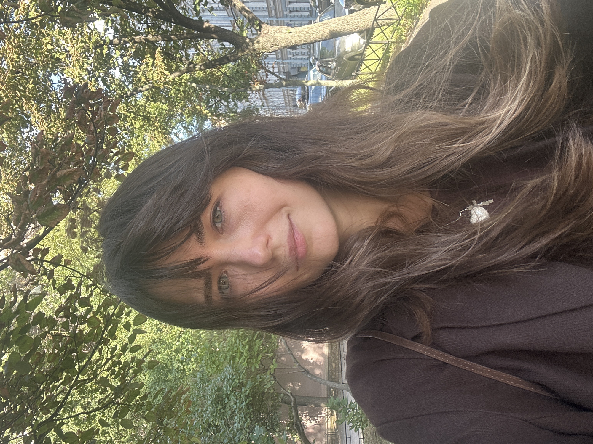 A medium shot, outdoor, selfie-style photo features a young woman with long, dark brown hair and bangs. She is wearing a dark brown jacket over a darker shirt and a silver necklace with a heart and cross pendant. Her expression is neutral, and she is looking directly at the camera. The background is slightly blurred, showing a park-like setting with trees and foliage. Sunlight casts shadows and highlights on the scene, suggesting it is daytime. In the distance, through the trees and a fence, a street with parked cars and buildings is visible, indicative of an urban environment. The architecture of the buildings suggests a European city, consistent with the city of Saint Petersburg, Russia. The overall atmosphere is casual and relaxed.