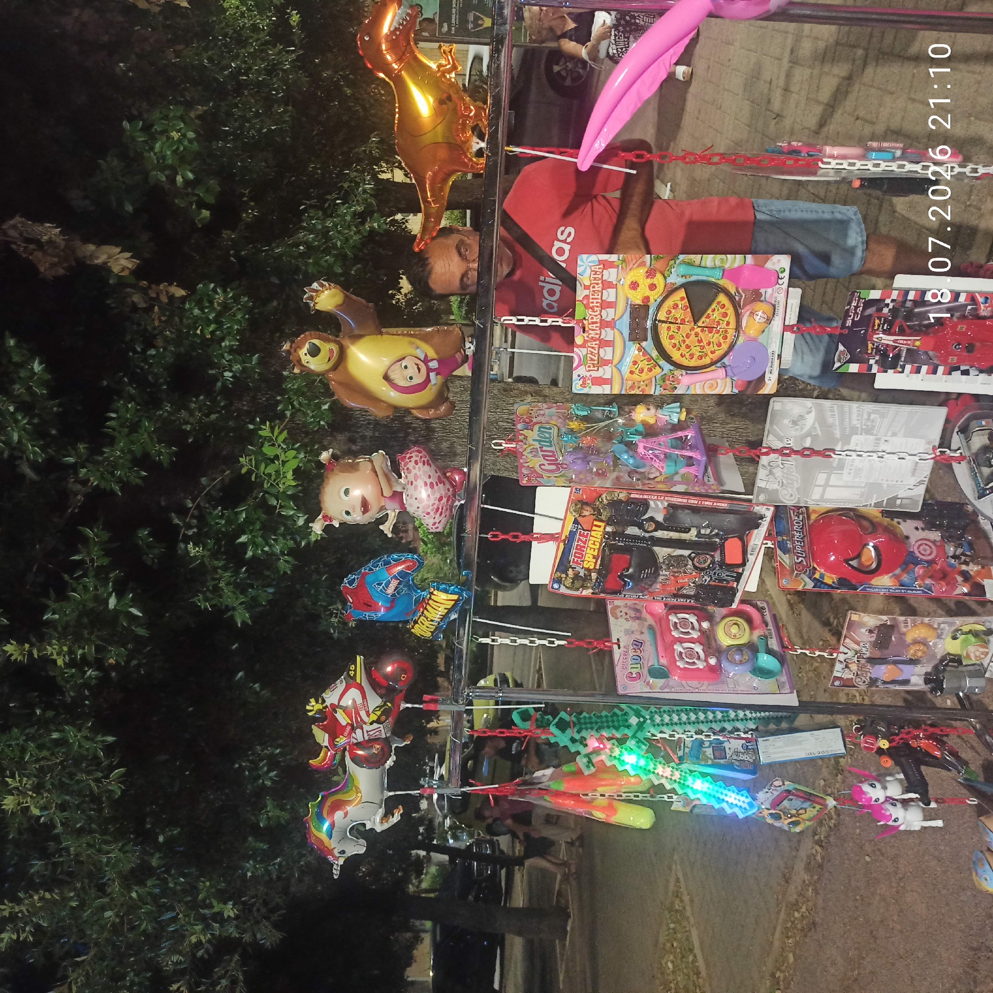 The image depicts a street stall in Bitonto, Italy, at night. The stall is laden with colorful balloons and various toys. In the background, trees and parked cars are visible, suggesting an urban setting. A man in a red t-shirt stands behind the stall, interacting with the merchandise. The overall atmosphere is that of a casual evening market.