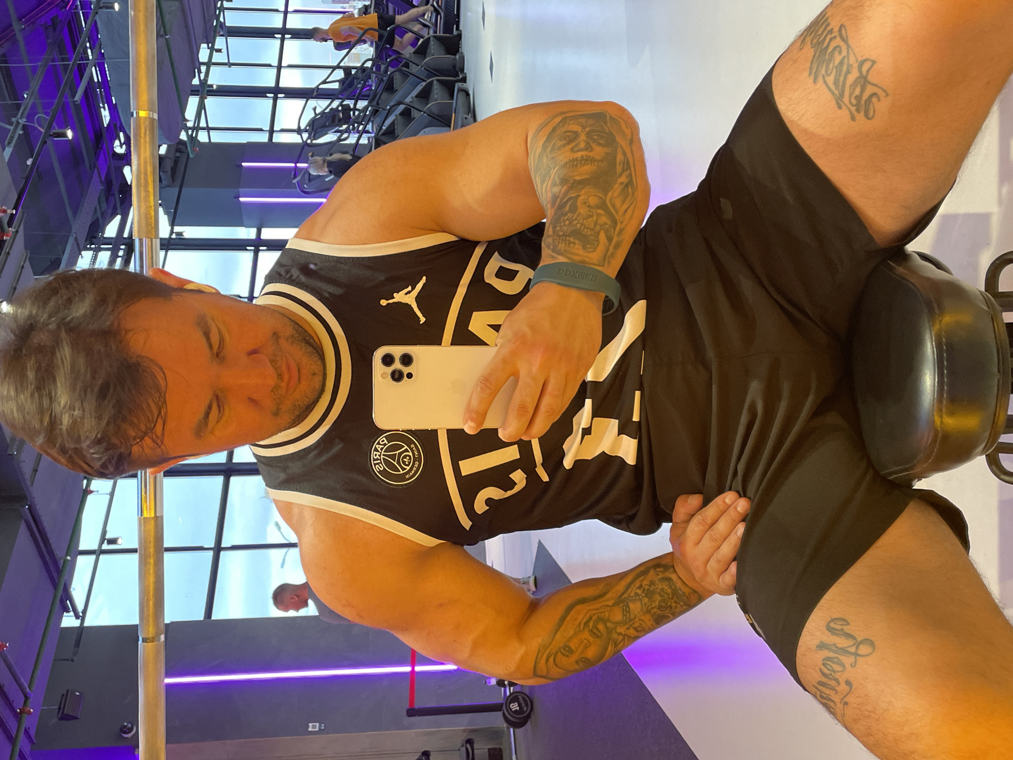 A medium shot, rotated to the left, features a fair-skinned, muscular man sitting on an exercise machine, likely in a gym, holding a white smartphone. The man is wearing a black and white basketball jersey with a Paris Saint-Germain logo and black shorts. He has tattoos on both arms and both thighs. The background includes gym equipment, including stationary bikes, and a window revealing a cityscape. The gym has a modern design, with white walls, purple accents, and a mirrored ceiling. Given the architectural details and the presence of a French soccer team's logo, the location is likely not in Kazan, Russia.