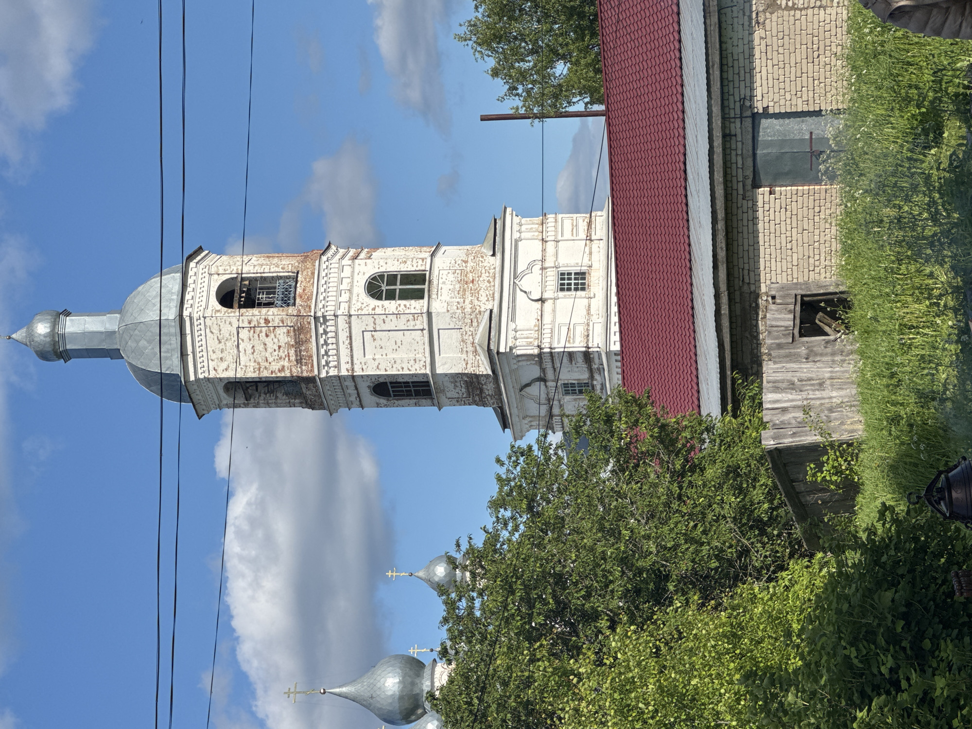 This media file depicts a serene, sunlit scene featuring a prominent religious structure in Луптюг, Russia. The main subject is a tall, weathered bell tower or church spire, constructed from white-painted brick with areas where the original red brick shows through. It has multiple levels, featuring both arched and rectangular windows, and is topped with a grey, metallic onion dome and a spire. Black power lines stretch across the upper part of the image, partially obscuring the tower and the sky.
To the right of the tower, a building with a distinct red corrugated metal roof is visible, suggesting an adjacent structure within the complex. Below it, there's a section of a light-colored brick wall with a metal covering, and an old wooden structure, possibly an outbuilding or shed, surrounded by overgrown green grass and wild plants. Lush green trees and bushes are scattered throughout the midground and foreground, partially obscuring other elements. In the lower left, behind some trees, several other grey, metallic church domes, each topped with a cross, are visible, indicating that the bell tower is part of a larger church or monastery complex.
The sky is a vibrant blue with scattered white, fluffy clouds, suggesting a clear and sunny daytime. There are no people visible, and no discernible text. The image's content is characteristic of traditional Russian rural or small-town architecture and natural landscapes. The entire image is presented as if rotated 90 degrees clockwise from its natural upright orientation.