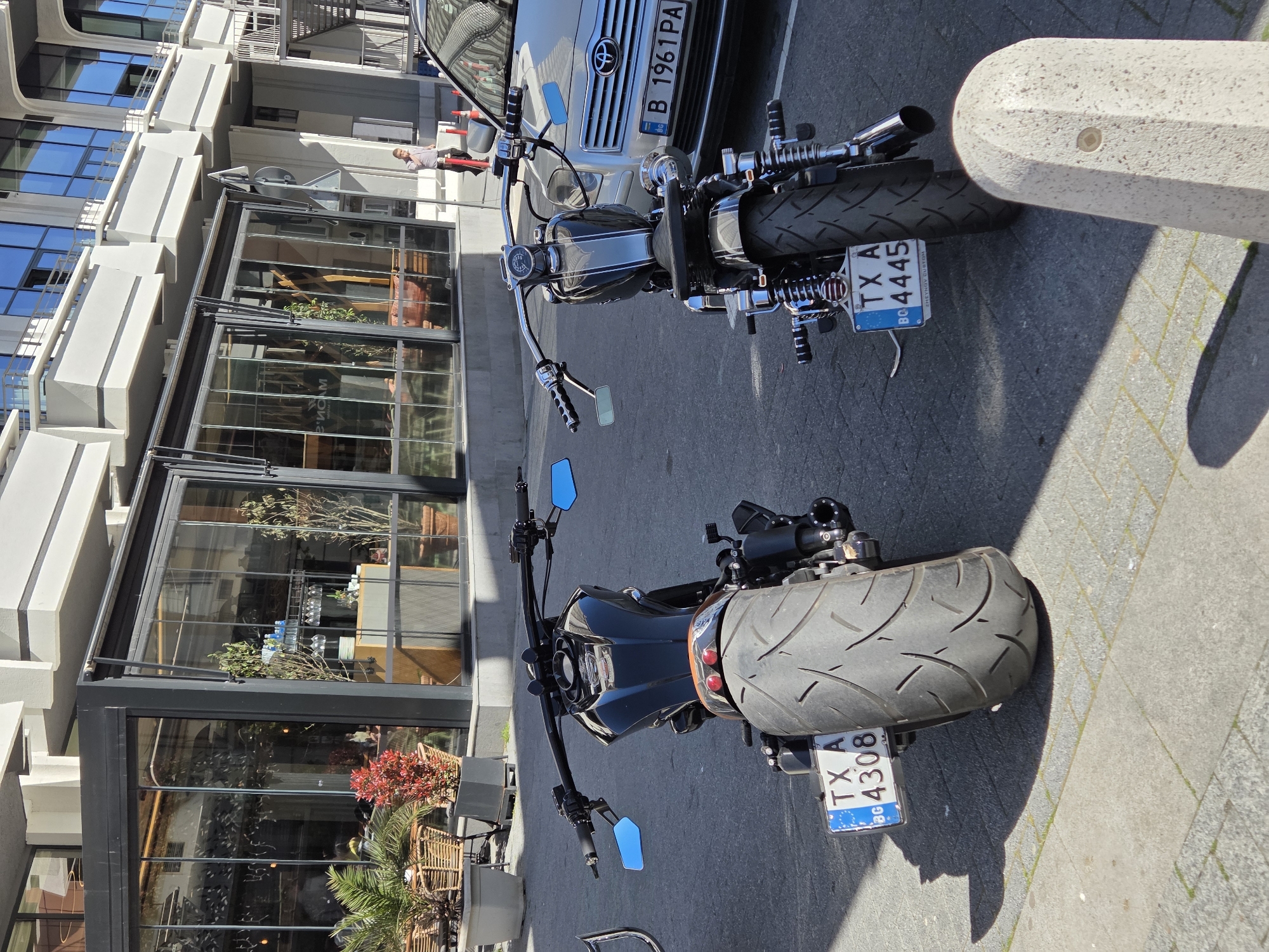Two motorcycles are parked on a grey pavement in front of a building with large glass windows. The first motorcycle, closer to the camera, is black with a license plate "TX A 4308 BC" visible. The second motorcycle is partially obscured by a silver car with license plate "B 1961 PA". The building appears to be a cafe or restaurant with visible plants and furniture inside and outside. A pedestrian is visible in the background on the left side. The city is Varna, Bulgaria.