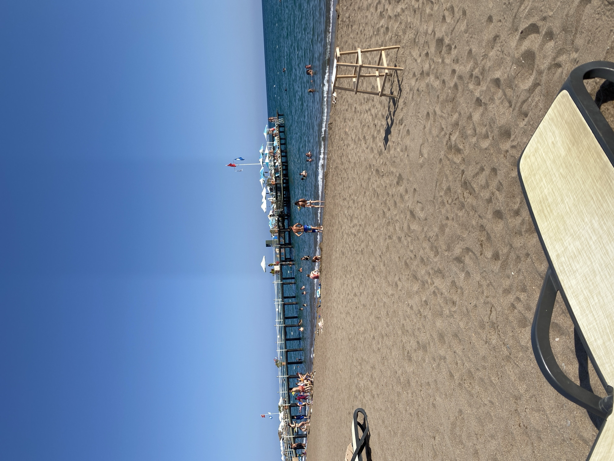 This is a wide-angle, eye-level shot of a crowded beach in Aksu, Turkey, on a sunny day. The foreground is dominated by a sandy beach with two lounge chairs, one in the lower left corner and another in the lower right. A wooden lifeguard chair is positioned in the middle ground, casting a shadow on the sand.
In the background, a long pier extends into the turquoise sea, lined with white umbrellas and people sunbathing. Numerous individuals are visible in the shallow water near the pier, swimming and wading. Some people are also walking along the pier and on the beach. The sky is a clear, bright blue. The overall impression is a lively and active beach scene.