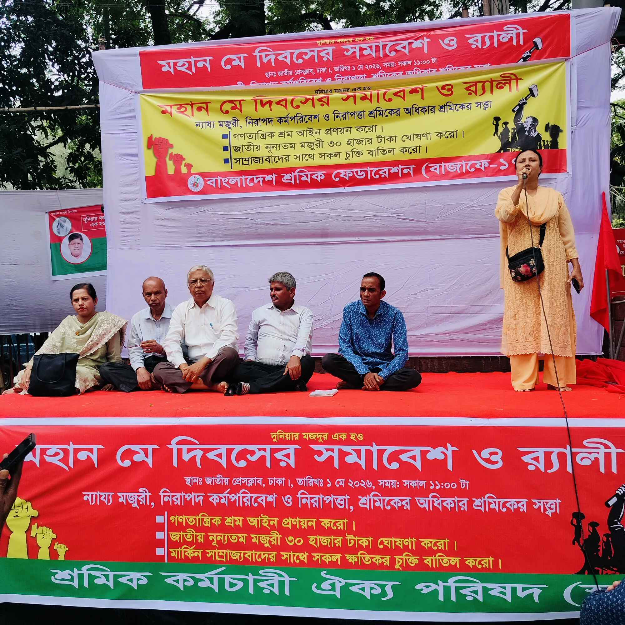 Here's a detailed description of the image:
**Scene:** An outdoor gathering, possibly a rally or demonstration, is taking place in Dhaka, Bangladesh. The location is identified as Jatiya Press Club, Dhaka.
**People:**
* A woman is standing at a microphone, seemingly addressing the crowd. She wears a light orange or yellow salwar kameez and a purse.
* Five other people are sitting cross-legged on a red platform or stage. There are three men and one woman sitting next to each other in the middle, and a fifth man on the other side.
* Hands holding a smartphone are partially visible on the left side of the image.
**Objects:**
* Two large banners or backdrops are visible. They promote the "মহান মে দিবস সমাবেশ ও র্যালী" (Great May Day Rally). The banners have demands related to workers' rights, including fair wages, safe working conditions, and a national minimum wage.
* A red flag stands on the right of the speaker.
* A microphone is placed in front of the speaker.
* A black purse is by one of the seated women.
**Environment:**
* The event appears to be held outdoors, with trees visible in the background.
* The red platform or stage provides a focal point for the speakers and seated participants.
**Actions & Interactions:**
* The woman is speaking into the microphone.
* The people sitting appear to be listening.
* The person holding the smartphone is likely recording or taking a picture of the event.
**Subjects:** The rally is centered on May Day (International Workers' Day) and advocates for worker's rights.
The banners contain the following information:
* Event: মহান মে দিবসের সমাবেশ ও র্যালী (Great May Day Rally)
* Location: জাতীয় প্রেসক্লাব, ঢাকা (National Press Club, Dhaka)
* Date: ১ মে ২০২৬ (May 1, 2026)
* Time: সকাল ১১:০০ টা (11:00 AM)
* Demands: ন্যায্য মজুরী, নিরাপদ কর্মপরিবেশ ও নিরাপত্তা, শ্রমিকের অধিকার শ্রমিকের সত্ত্বা (Fair wages, safe working conditions and safety, workers' rights, workers' power), গণতান্ত্রিক শ্রম আইন প্রণয়ন করো (Enact democratic labor laws), জাতীয় নূন্যতম মজুরী ৩০ হাজার টাকা ঘোষণা করো (Declare a national minimum wage of 30,000 Taka), সাম্রাজ্যবাদের সাথে সকল চুক্তি বাতিল করো (Cancel all treaties with imperialism).
**Note:** This description is based solely on the provided image.