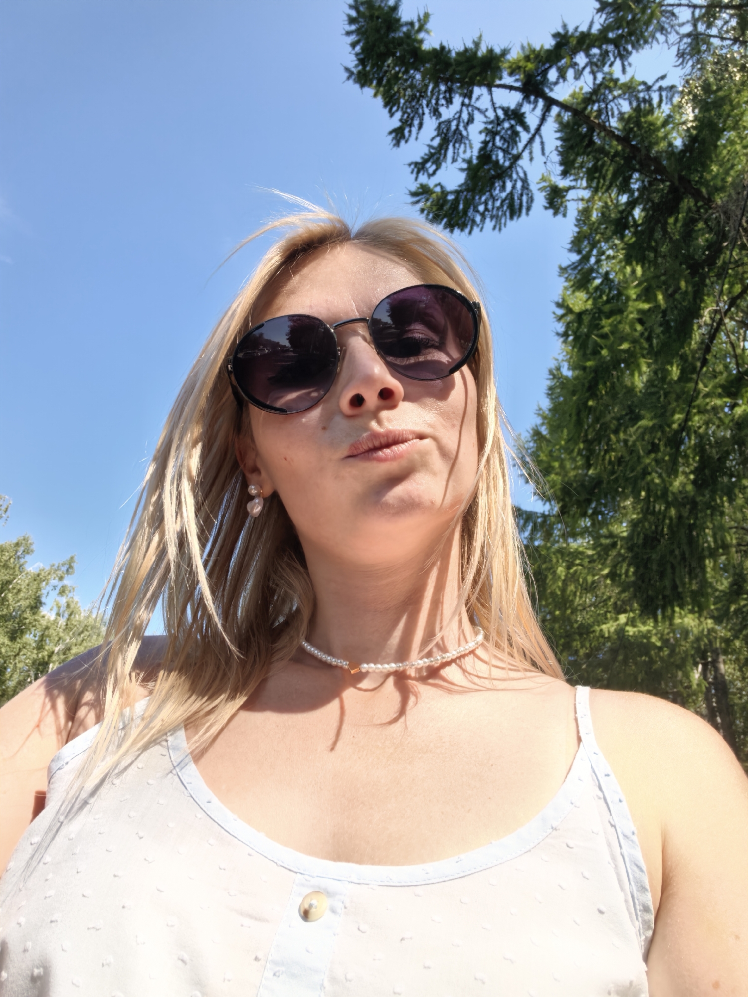 The image is a close-up, low-angle shot of a fair-skinned woman with blonde hair and sunglasses. She is wearing a light-colored camisole and a pearl necklace. The background consists of a clear blue sky and green trees, suggesting an outdoor setting. There are no visible landmarks or indicators to place this scene specifically within the city of Sterlitamak, Russia.