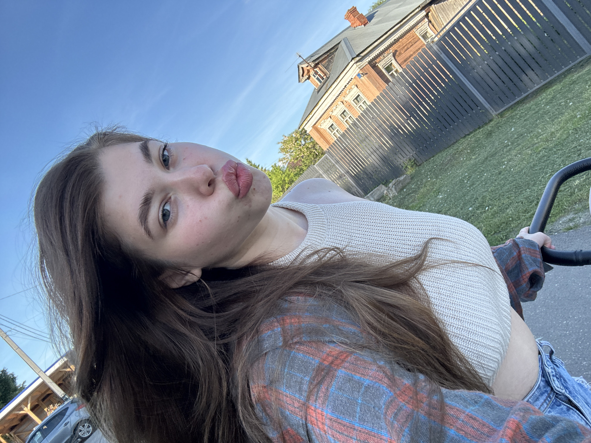 A young woman with long brown hair, likely in her late teens or early twenties, poses for a selfie outdoors. She is looking directly at the camera with a playful "kissy face" expression. Her attire includes a light-colored, possibly beige or off-white, ribbed crop top, and a plaid shirt with red, blue, and grey checks worn loosely or tied around her waist. She also wears blue denim jeans. Her left hand is visible, holding a black handle, possibly from a stroller or bicycle.
The background features a clear blue sky, suggesting bright daylight, possibly late afternoon or early evening. To the right, a traditional Russian-style wooden house with ornate carvings, a dark grey roof, and a red brick chimney stands behind a dark wooden slatted fence. The fence runs along a strip of green grass and a paved path or road. In the far background on the left, a utility pole, power lines, and a silver car are partially visible. The setting appears to be a casual residential or semi-rural area, consistent with Мещёры, Russia. The overall mood is lighthearted and casual. There is no visible text in the image.