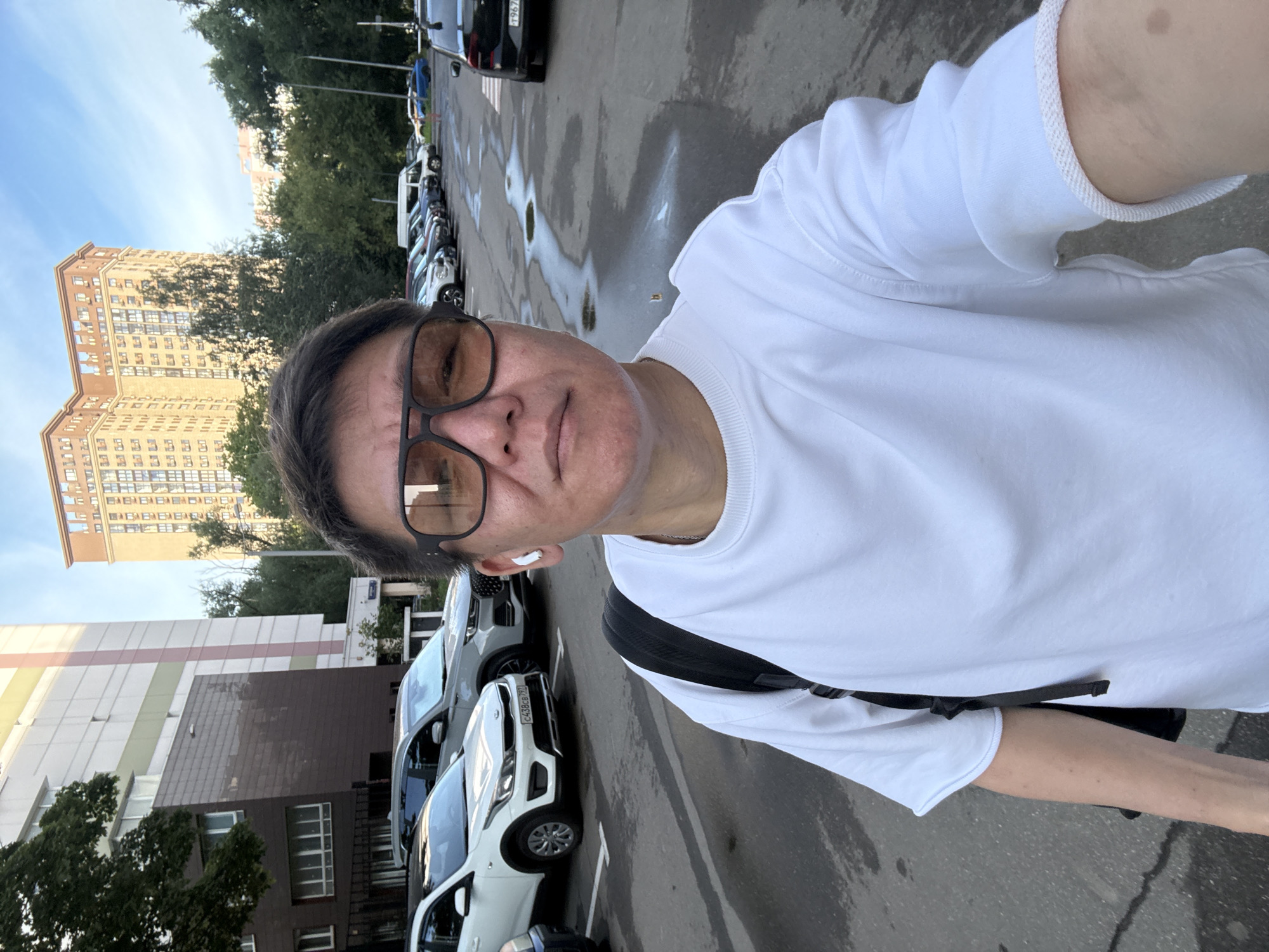 A person is taking a selfie in an outdoor parking lot in Moscow, Russia. The person is wearing a white sweatshirt and dark-rimmed sunglasses. They have white earbuds in their ears and a black backpack is visible on their shoulder. The parking lot is paved and there are several cars parked in it. In the background, there are apartment buildings and trees. The sky is blue with some clouds.