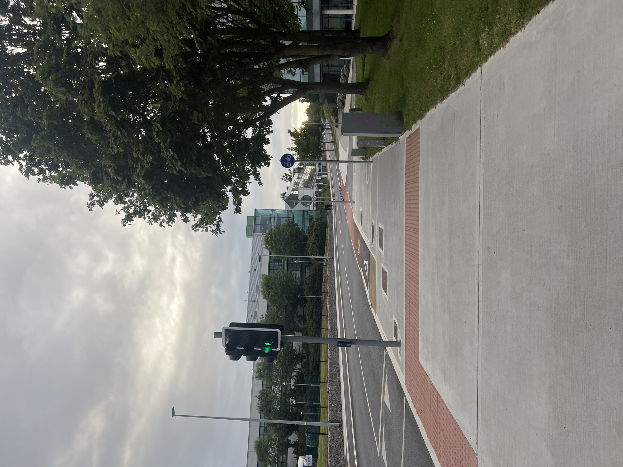 The image depicts a street scene in Balbriggan, Ireland, likely during overcast daylight. In the foreground, a concrete sidewalk runs alongside a paved road. The sidewalk has a textured red strip at its edge. To the right of the sidewalk is a grassy area with several mature trees.
On the sidewalk, there are several gray concrete bollards and a utility box. A traffic light is visible on a pole, displaying a green light. Next to the traffic light, a blue sign with a white bicycle symbol indicates a cycle path. The road is marked with white lines, and further down, red markings are visible, possibly indicating a cycle lane.
In the background, several modern buildings with large glass windows and trees are visible. The sky is overcast with diffused sunlight. The scene appears to be an urban or suburban environment with dedicated pedestrian and cycle infrastructure. There are no people clearly visible in the immediate foreground or midground of the image.