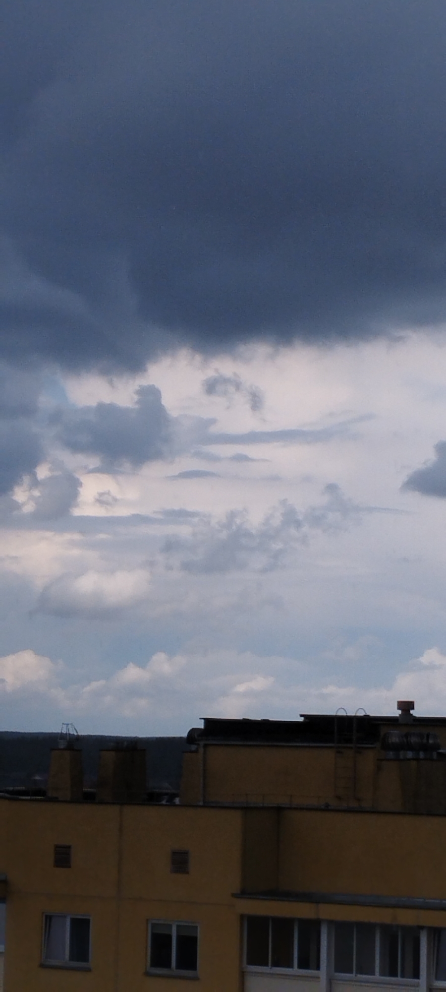 A low-angle, vertical, full shot captures a building and a stormy sky above in Skidzieĺ, Belarus. The sky dominates the upper two-thirds of the frame, filled with dark, heavy clouds that suggest an impending storm. Lighter clouds break through in the middle ground, creating a textured, layered effect. The lower third of the image features the rooftop and upper floors of a yellow building. The building's facade is segmented by horizontal lines, with visible windows and ventilation grates. Several rooftop structures are present, including what appears to be a ladder leading to a higher level, exhaust pipes, and other utility equipment. The overall impression is one of a moody, overcast day over an urban landscape.