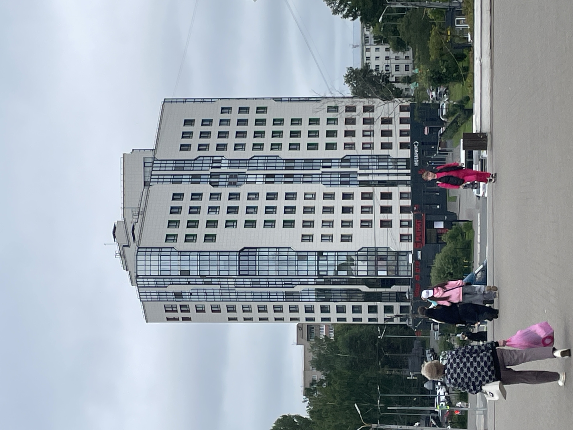 The image captures a modern, multi-story building with a predominantly white facade and a central section of dark-framed, floor-to-ceiling windows in the city of Kirov, Russia. The sky is overcast with a dull, grey hue. In the foreground, several individuals are walking on a paved surface. Closest to the viewer, an older woman with blonde hair, wearing a patterned jacket and grey trousers, carries a white handbag and a large pink shopping bag. Further back, a group of three people walk together; one is wearing a black hijab and a long dark coat, another a pink hooded jacket and grey trousers, and the third person is mostly obscured. To the right, a young person in a bright pink tracksuit walks towards the viewer, holding a dark bag. In the background, more buildings and trees are visible, along with parked cars and streetlights, suggesting an urban environment. A sign with Russian text, "НОТАРИУС", is visible on the lower part of the modern building. The overall scene depicts a typical day in an urban setting with people going about their business.