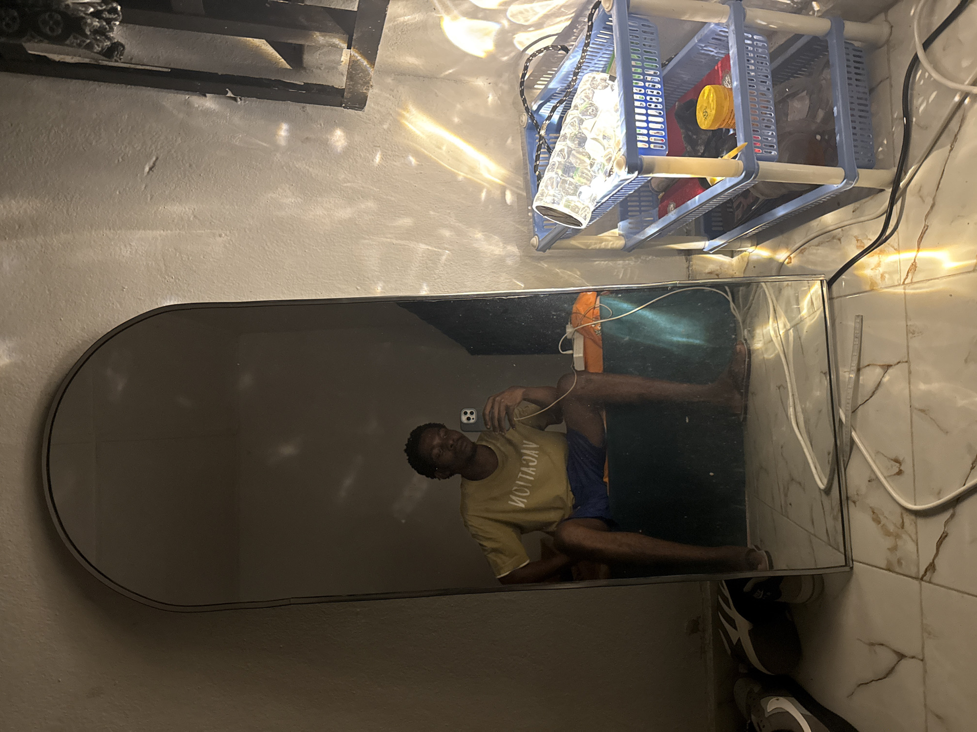 A person is taking a mirror selfie in a room. The person is wearing a yellow t-shirt with the word "VACATION" in white text and blue shorts. They are sitting on a dark green surface. In the background, a white wall is visible with light patterns from what appears to be a decorative lamp. To the right, a blue plastic shelf unit holds various items, including a clear decorative lamp and a yellow container. The floor is tiled with a marble pattern. Shoes are visible on the floor to the left of the mirror. The scene suggests a casual indoor setting. This appears to be a private residence in Uyo, Nigeria.