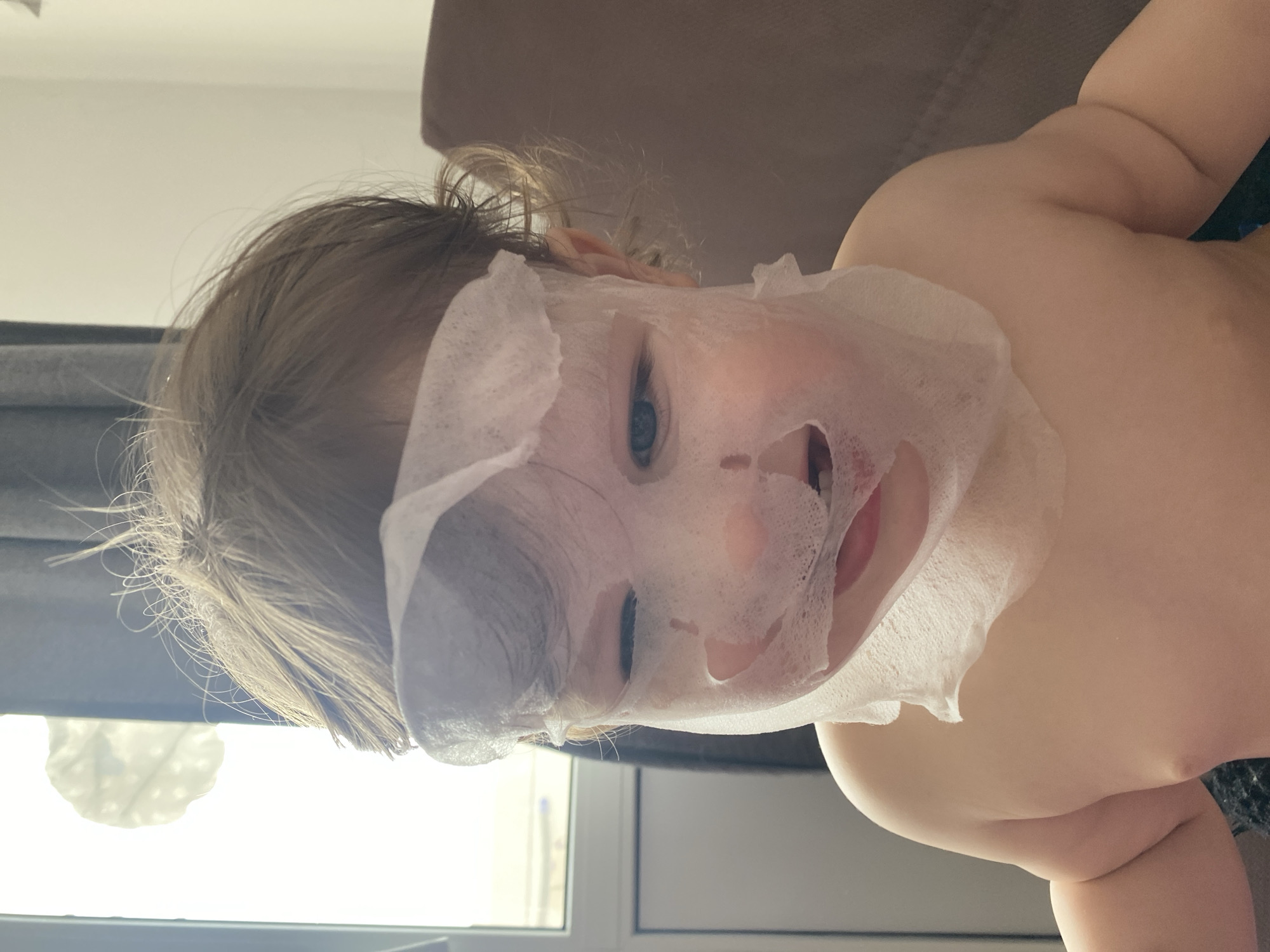 A close-up shot depicts a young child with a face mask applied. The child has fair skin and light-colored hair, with a section pulled up on the head. The face mask appears to be a thin, sheet-like material covering the majority of the face. The child is positioned indoors, with part of a window visible in the background along with some furniture. The location is unknown.