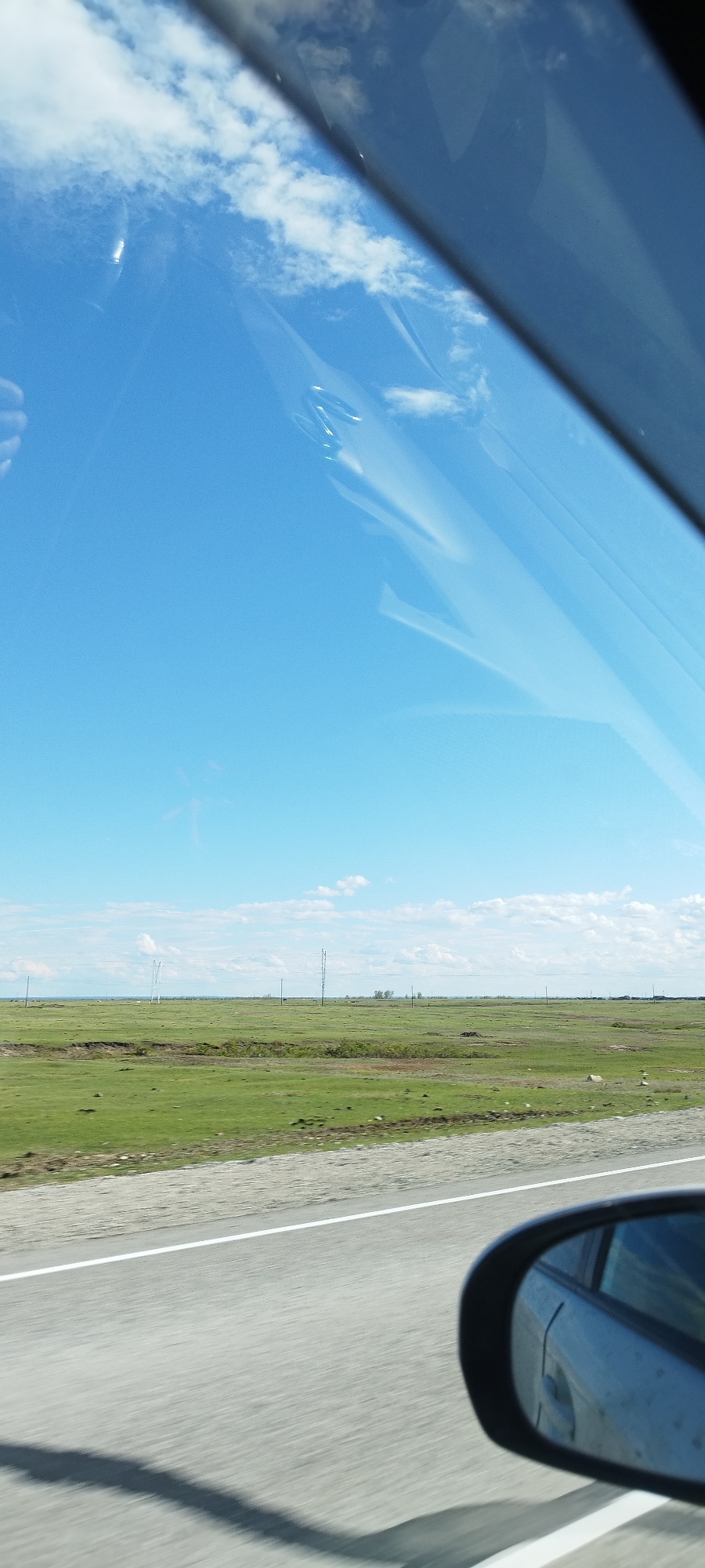 The image depicts a view from inside a moving vehicle, likely a car. The foreground consists of a portion of the vehicle's side mirror and a segment of the asphalt road with a white lane marker. Beyond the road, there is a vast, flat expanse of grassland stretching towards the horizon. Several utility poles are sparsely scattered across the landscape. The sky is a clear, bright blue with scattered white clouds. The location is the city of Ulakh-An, Russia.