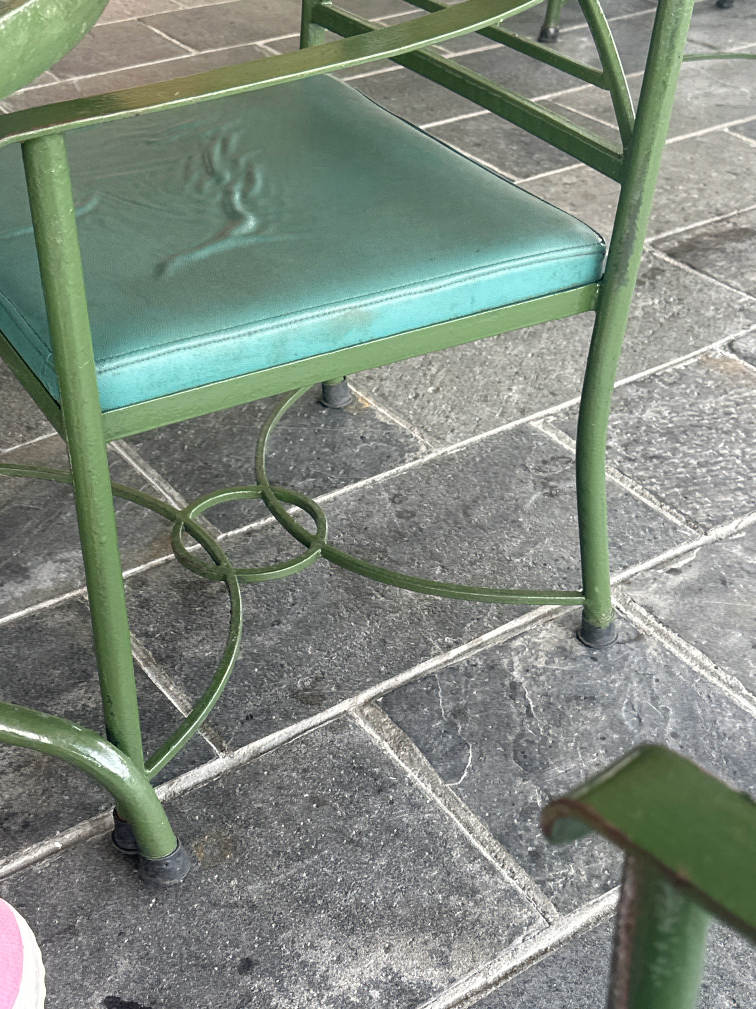 A close-up, eye-level shot focuses on the lower half of a weathered, green metal chair with a teal-colored seat cushion. The cushion appears slightly creased. The chair's legs rest on a stone-tiled patio. The tiles are dark gray with lighter grout lines. The chair has a decorative metal base with looped elements. In the bottom right corner, a portion of another green chair arm is visible, out of focus. No people are visible in the frame. The scene is devoid of specific environmental context that would place it in Kageshwori Manohara, Nepal; it could be any outdoor patio with similar furnishings and flooring.