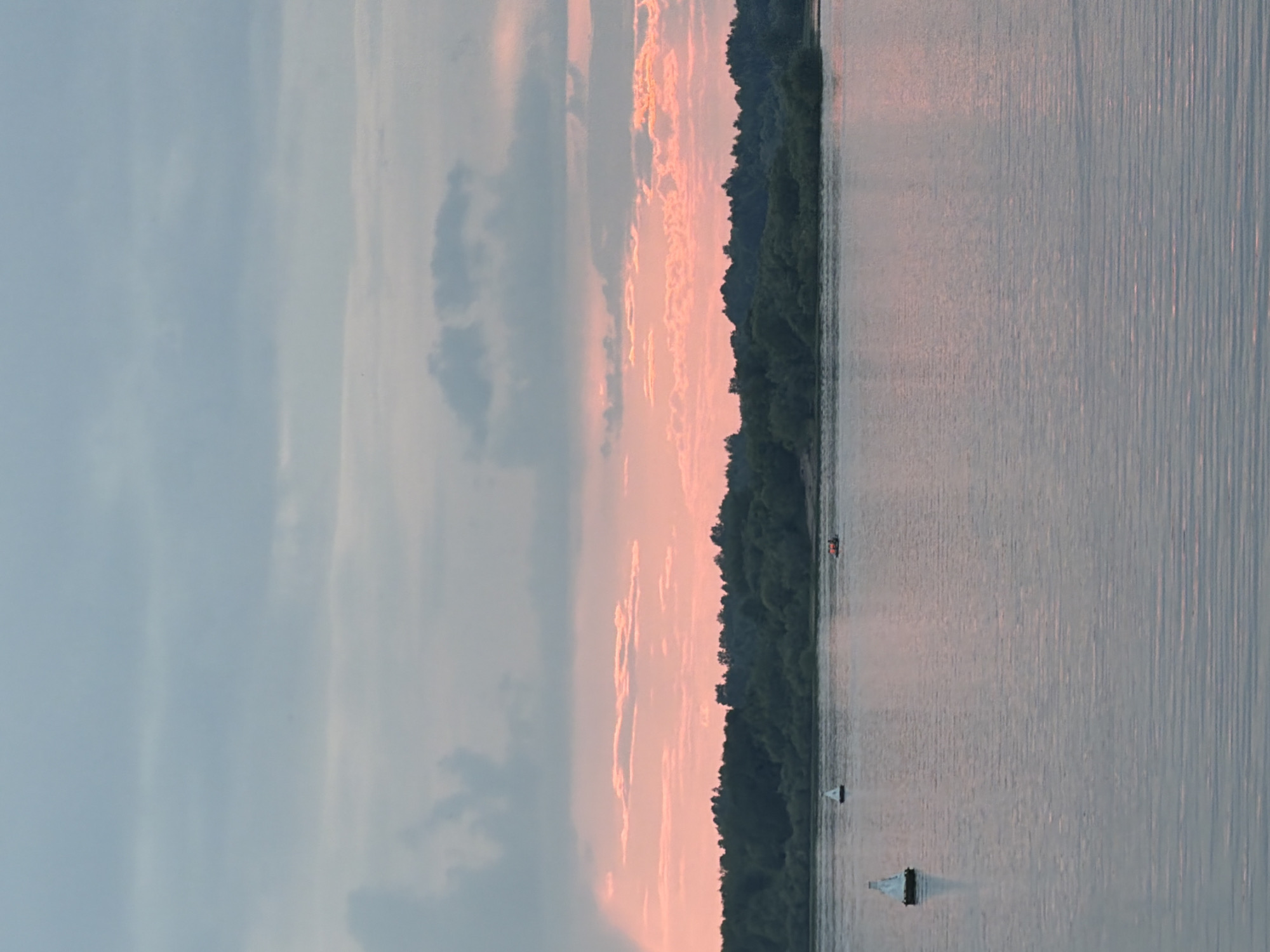 A wide, calm river occupies the lower two-thirds of the frame, reflecting the soft hues of the sky. The water is a muted silvery blue with subtle horizontal ripples. On the left side of the river, a white buoy with a black base floats. In the middle distance, another white buoy is visible. Further to the right, a small orange boat with what appears to be a person or two on board is moving across the water.
The opposite bank of the river is lined with a dense, dark green forest, its silhouette sharp against the sky. Above the treeline, the sky transitions through soft pinks and oranges near the horizon, indicating either sunrise or sunset. Higher in the sky, the clouds are a blend of pale blue and gray, with some areas appearing slightly darker and more textured. The overall atmosphere is serene and tranquil. The scene is consistent with a view along the Kama River in the vicinity of Solikamsk, Russia, during the twilight hours.