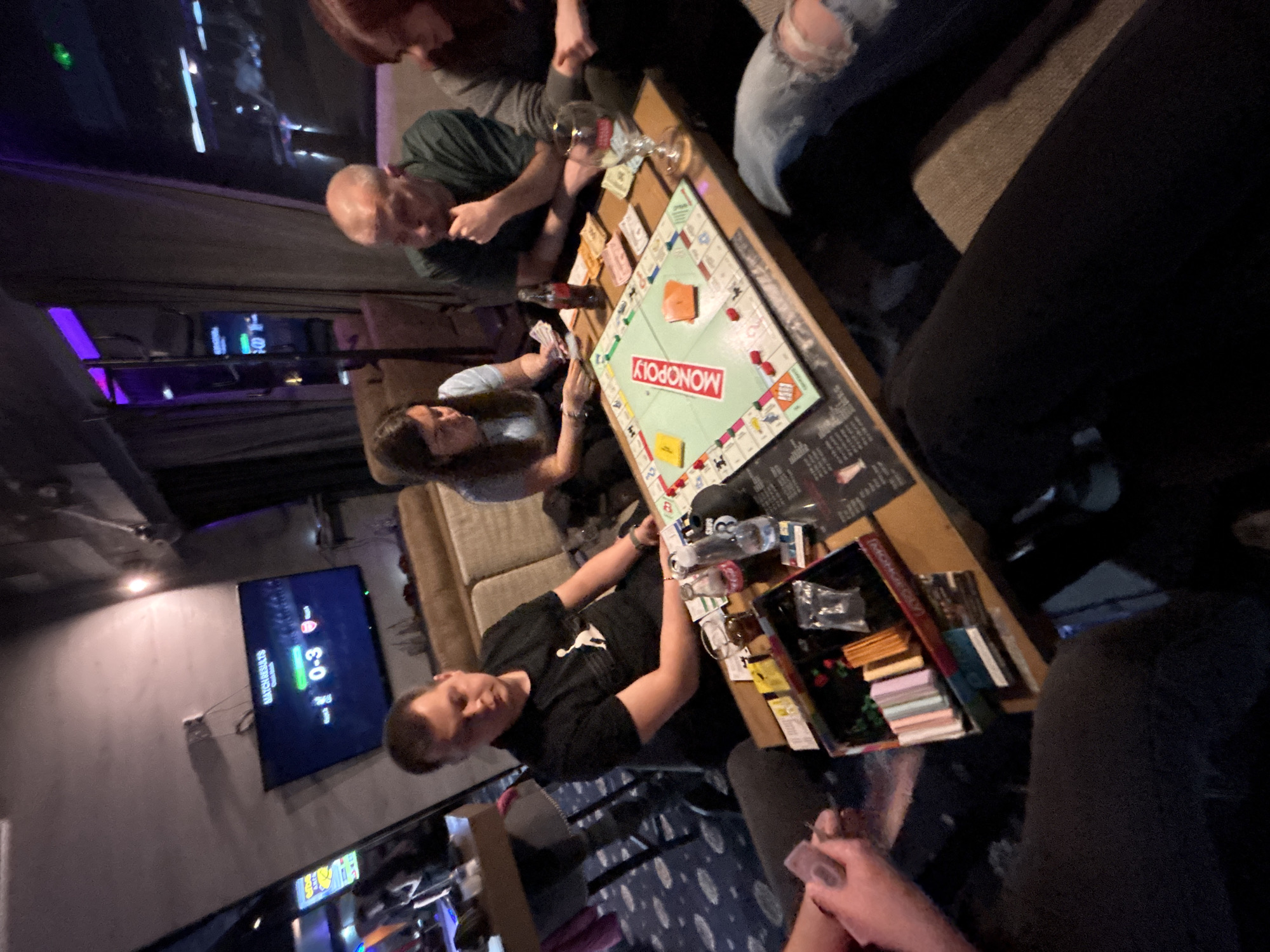 A group of people are playing Monopoly at a table in an indoor setting. The game is underway with money, cards, and pieces scattered on the board. There are at least four people seated around the table, some holding cards and others watching the game. The setting appears to be a lounge or recreational room, with a television showing a soccer match score in the background. Beyond the players, there is a bar-like area with various fixtures visible. The location, based on the information requested, is Plovdiv, Bulgaria.