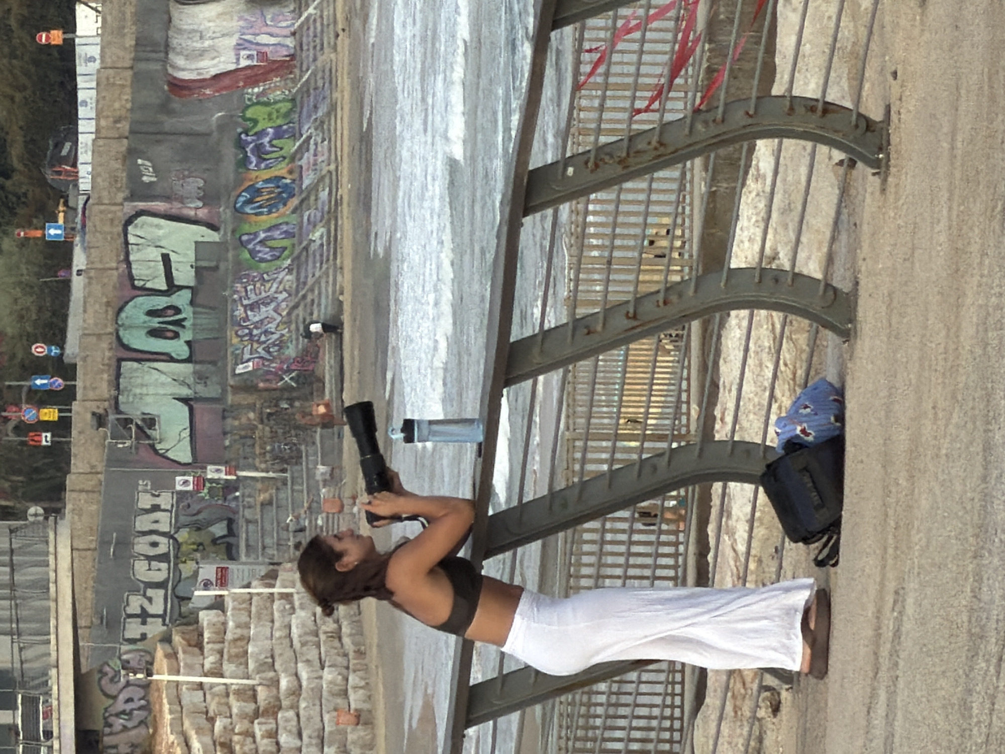 This media file captures a candid shot of a woman standing on a paved walkway next to a metal railing, with the sea and beach visible in the background. The image is oriented vertically, suggesting it may have been taken with a mobile phone. The woman is dressed in a dark sports bra and flowing white pants, with brown hair tied back. She is holding a large camera with a telephoto lens to her eye, appearing to be taking photographs.
Behind her, the walkway is lined with rough-hewn stone blocks, and further back, a wall is covered in vibrant graffiti art. The graffiti features various tags and images in bright colors. In the distance, the sea appears calm with gentle waves washing onto a sandy beach. The time of day seems to be late afternoon or early evening, given the soft lighting and slightly elongated shadows.
To the left of the frame, there are traffic signs and what looks like a street or promenade. On the right, near the woman, a dark backpack and a blue patterned item are placed on the ground next to the railing. The overall atmosphere appears relaxed and casual, suggesting a tourist or local enjoying a walk and taking in the scenery. There is no visible text that can be clearly deciphered in the image, other than the graffiti on the wall, which is not legible in this resolution.