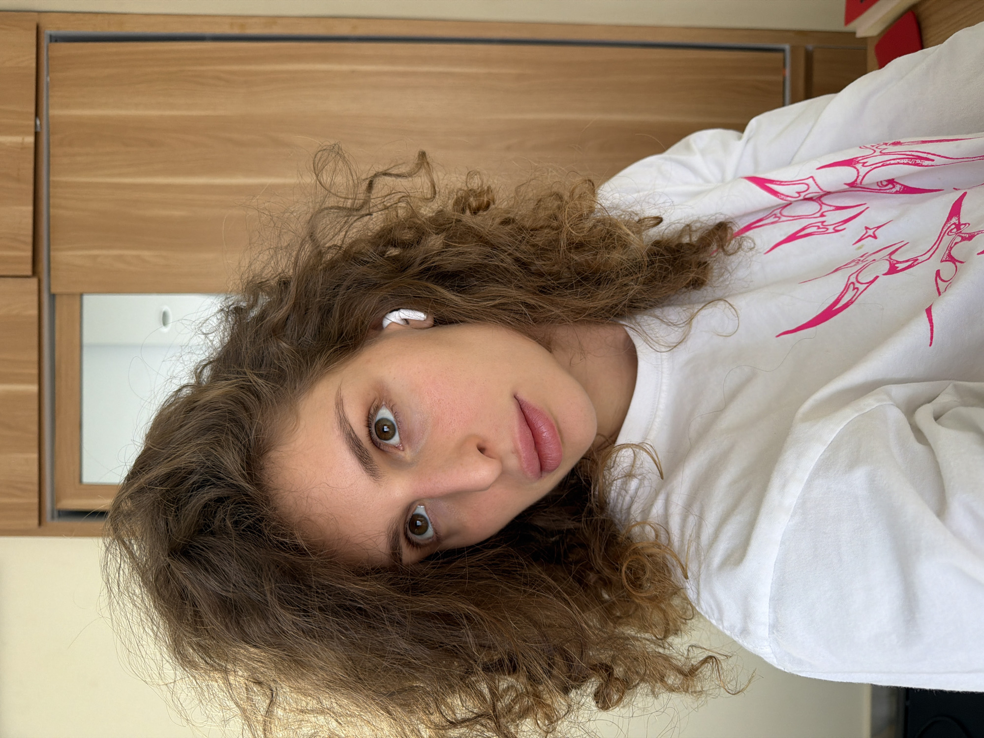 The scene is set in North Kuta, Indonesia. A young woman is depicted in a close-up, self-shot portrait. She possesses voluminous, medium-brown curly hair that frames her face and extends over her shoulders, appearing naturally textured with some stray strands. Her skin is fair, and her brown eyes are directed upwards and slightly to the left. Her full lips are slightly parted, revealing a natural pink hue. A white wireless earbud is visible in her right ear. She is wearing a white long-sleeved t-shirt or sweatshirt adorned with an intricate, spiky pink graphic design across the chest and right sleeve (from the viewer's perspective). The background consists of a plain light-colored wall on the left side of the frame and a modern wooden cabinet system on the right. The wooden system features vertical paneling and incorporates a mirror that reflects a light ceiling with a single recessed light fixture. A small, partially visible red object rests on a surface in the far lower right corner of the image.