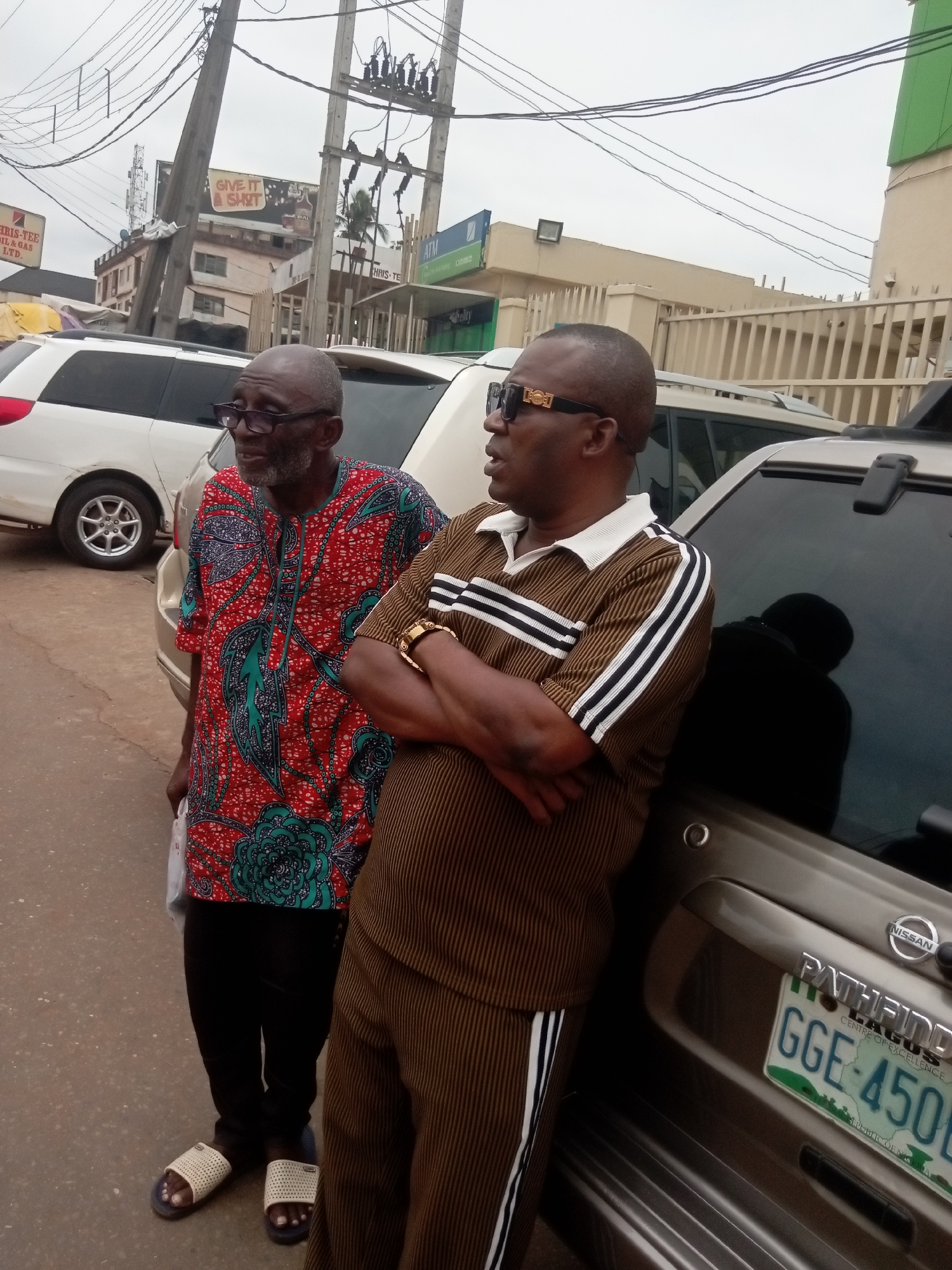 Two African men stand beside a silver Nissan Pathfinder with license plate GGE-450. The man on the left wears a red and teal patterned shirt, black pants, and sandals. The man on the right is leaning against the vehicle with his arms crossed, wearing a brown tracksuit with white stripes and sunglasses. In the background are buildings with signs that read "CHRIS-TEE OIL & GAS LTD" and "GIVE IT A SHOT." Overhead are numerous power lines. The license plate indicates Lagos, Nigeria, but the text specifies Nkpor as the location.