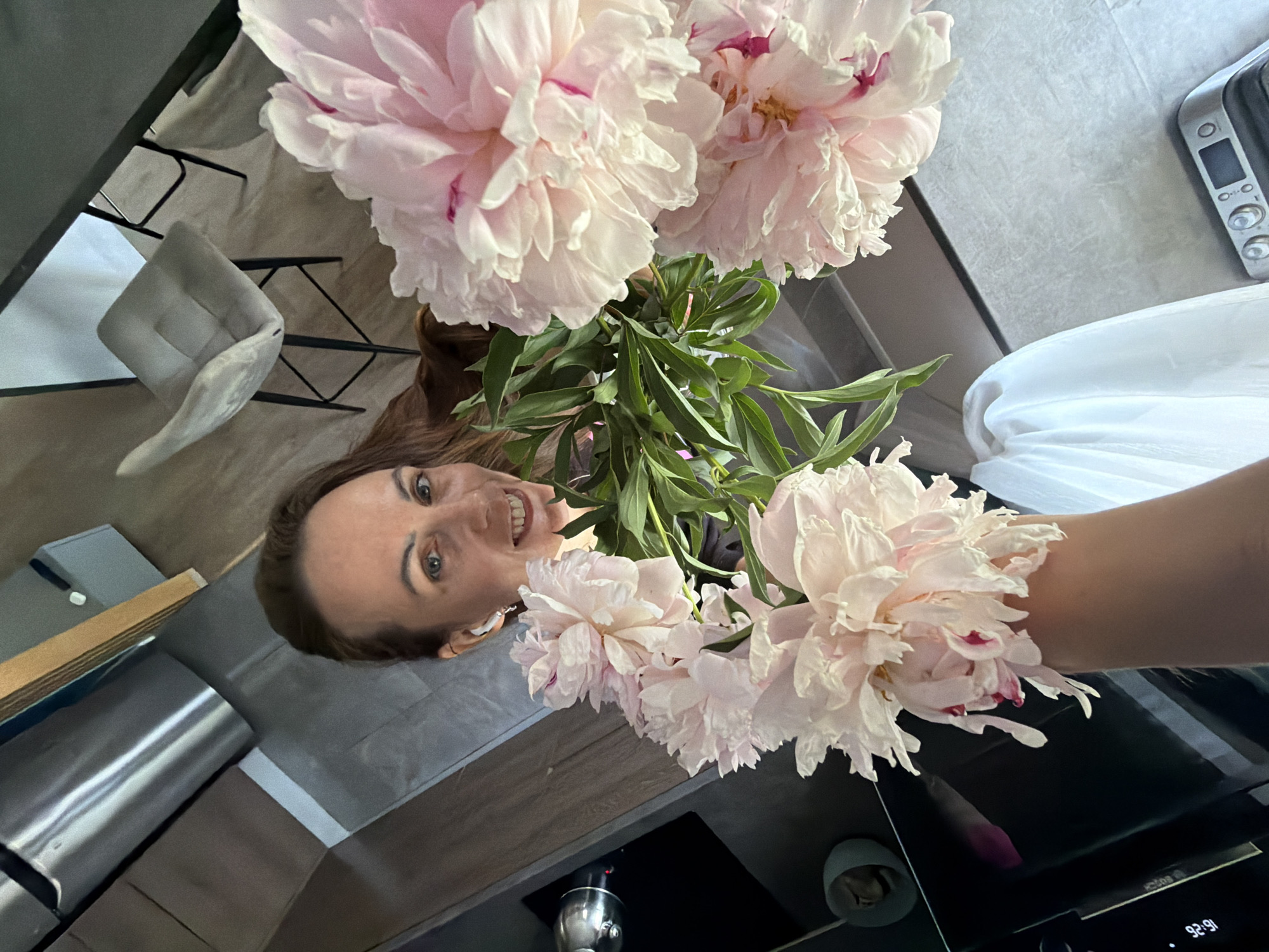 A woman is holding a bouquet of peonies and taking a selfie in a modern kitchen. She is smiling and has on white wireless earbuds. In the background, there are stainless steel appliances and chairs. The image is taken from a high angle, looking down at the woman and the flowers.