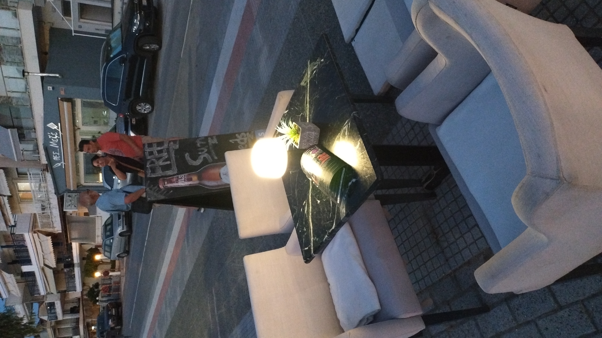 In Neoi Epivates, Greece, an outdoor seating area of a restaurant is depicted. Three upholstered chairs surround a dark marble-topped table. A small, illuminated lamp and a potted plant are on the table, along with a menu or magazine. A chalk sign with "FREE" and other writing is propped nearby. In the background, several people are visible, including a man in a blue shirt and two people in casual attire near a building with awnings. Cars are parked along the street, and a building with multiple floors is to the left. The ground is paved with cobblestones and concrete. The lighting suggests it is dusk or evening.