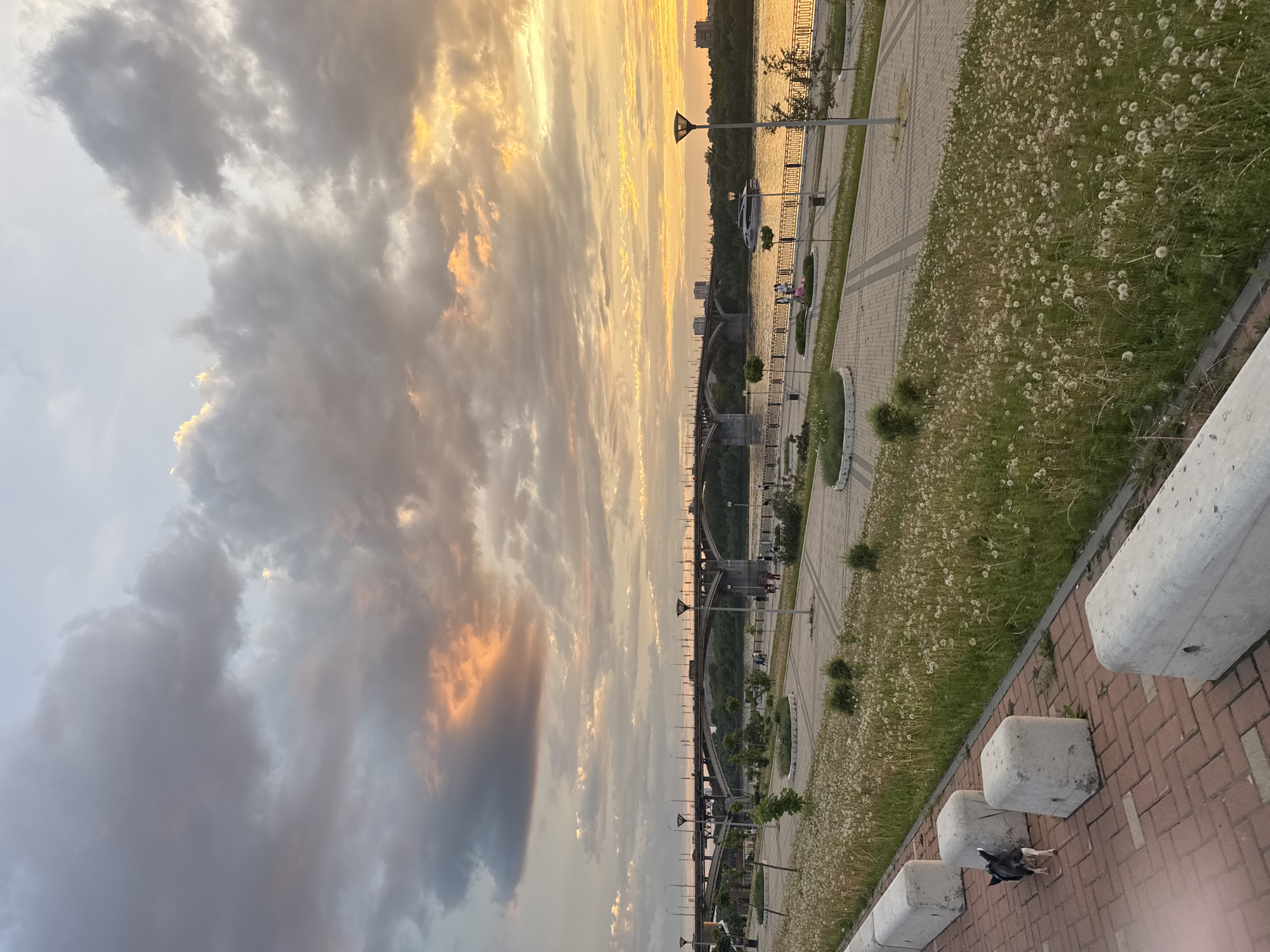 The image depicts a sunset scene in Nizhny Novgorod, Russia. The sky is filled with dramatic clouds, some tinged with gold from the setting sun. In the distance, a bridge stretches across a body of water, likely the Volga River. Along the riverbank, there's a paved walkway with lampposts and some greenery. There are some people visible, including a family on the walkway. In the foreground, there's a low brick wall and some concrete barriers. A bird is visible near the barriers, foraging on the ground. The general setting is an urban waterfront at dusk.