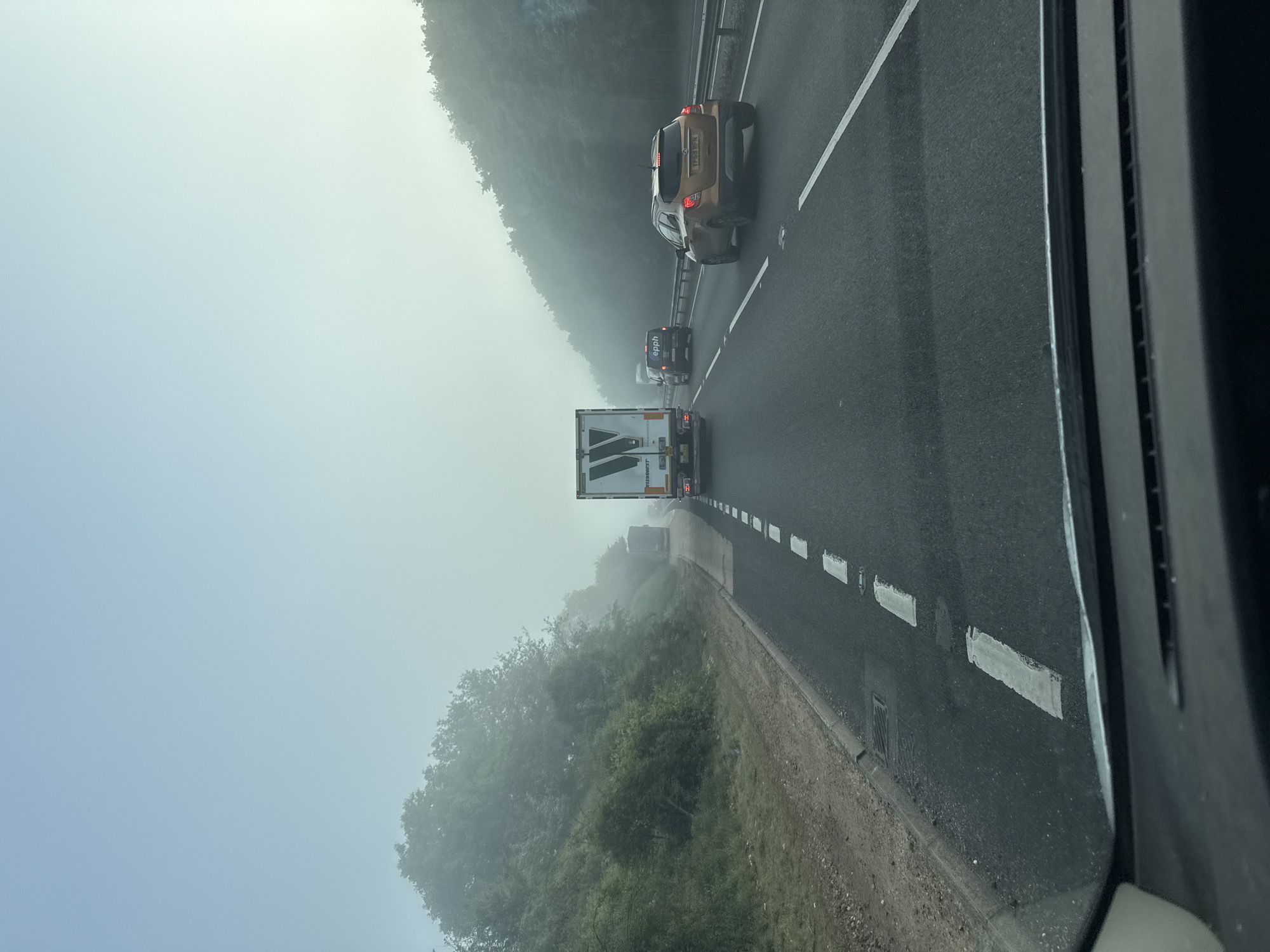 The image shows a foggy highway scene in Babergh, United Kingdom. Several vehicles, including a large truck and a gold SUV, are visible on the multi-lane road. The environment is obscured by heavy fog, reducing visibility. Trees line the side of the road. The photo appears to be taken from inside a vehicle.