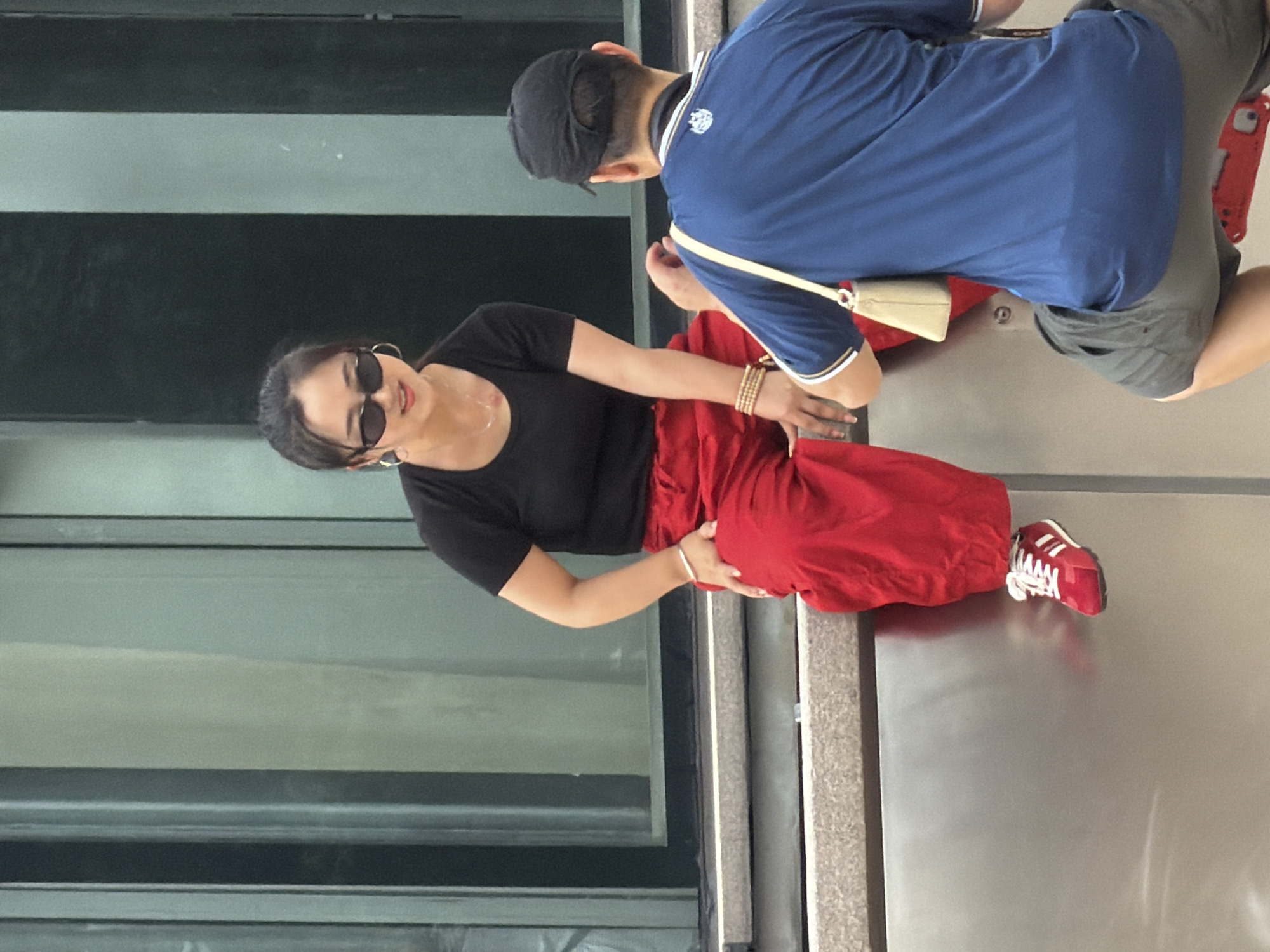 A young Asian woman with dark hair pulled back and wearing black sunglasses, a black t-shirt, and bright red pants is sitting on a ledge. She is facing a person from behind, who is wearing a blue t-shirt and a black cap. The woman has a necklace and bracelets on her wrists. Her red sneakers are visible. The man has a light-colored bag slung over his shoulder. They are in an urban environment, with a building in the background. The scene appears to be in Yuzhong District, China.