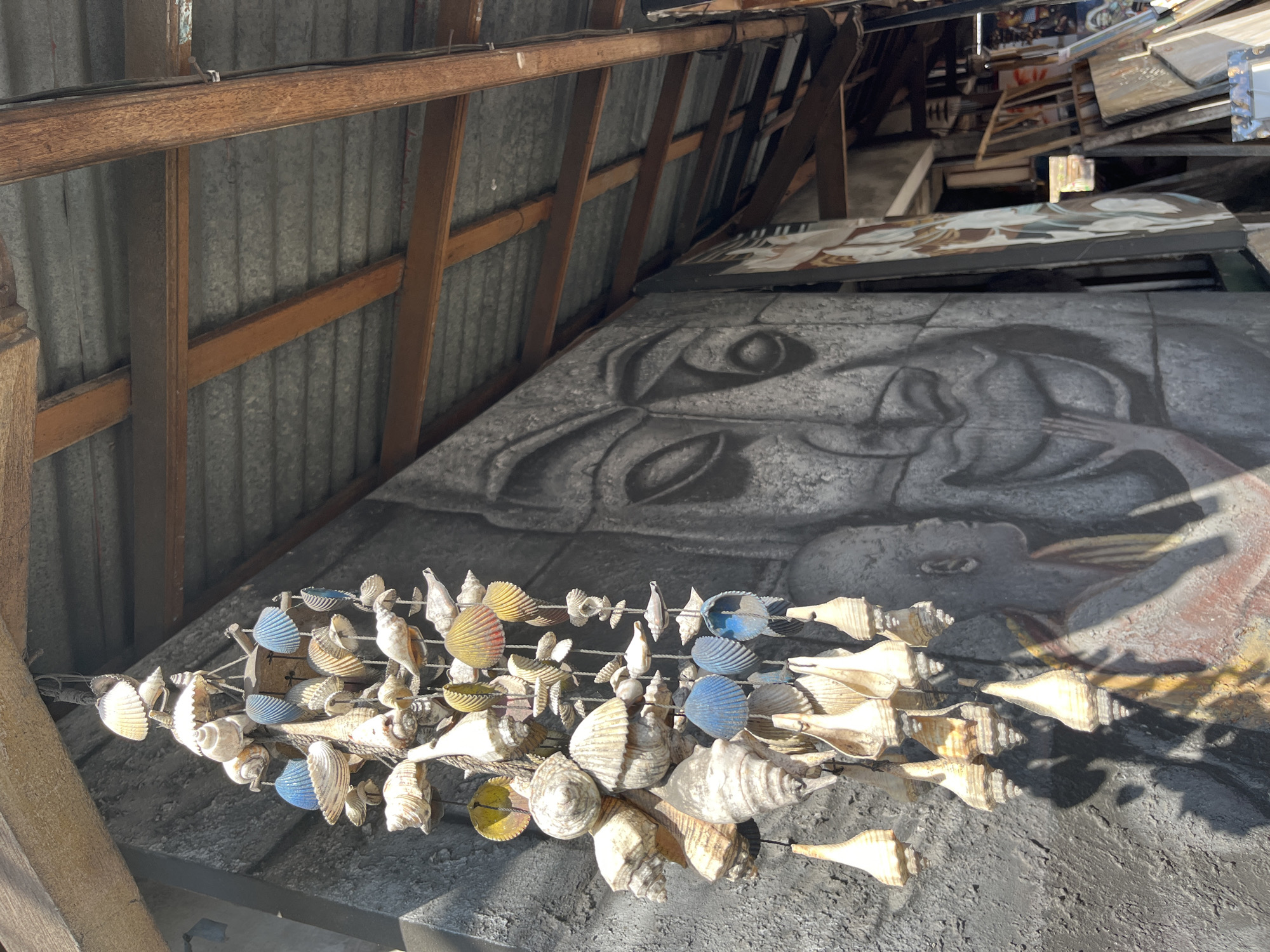 A seashell mobile hangs from the wooden eaves of a corrugated metal roofed structure. The mobile is composed of numerous seashells of varying sizes and shapes, some painted blue or yellow, strung together with twine. In the background, a large mural is painted on a textured dark gray wall, depicting a stylized face with closed eyes and a hand touching its chin. To the right of the mural, various canvases and artworks are leaned against the wall and stacked on the floor, suggesting an artist's studio or storage area. The overall scene appears to be outdoors, possibly in a market or workshop setting, in Sukawati, Indonesia. The lighting suggests a bright, sunny day. No people are visible in the image.