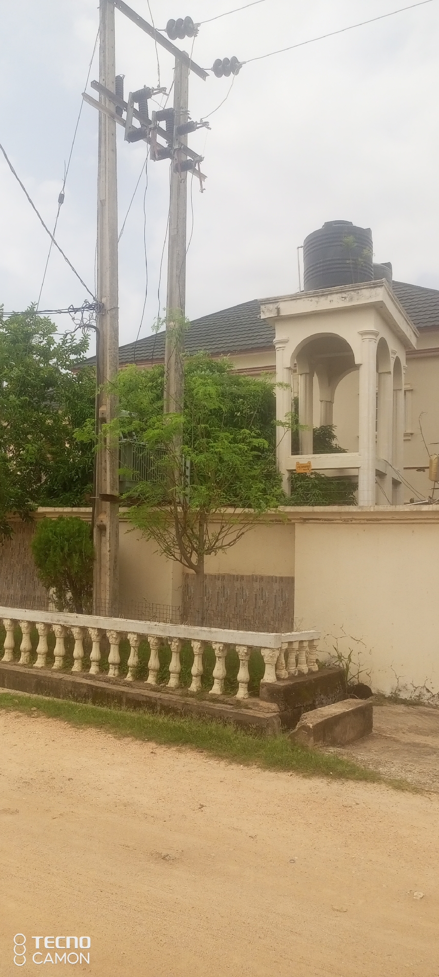 The image depicts a residential area in Jalingo, Nigeria, under an overcast sky. In the foreground, a dirt road or pathway transitions to a patch of grass. A low, decorative white balustrade with intricately carved posts runs along a concrete border. Behind the balustrade, a beige-colored wall separates the property from the street.
Dominating the mid-ground are two large, weathered utility poles supporting a complex network of electrical wires and insulators. Dense green foliage from various trees and shrubs surrounds the base of the poles and extends towards the right side of the image. Partially obscured by the greenery is a light beige house with a dark, tiled roof. An arched, open-air portico with white columns is visible on the front of the house. A large black water tank sits atop the roof. The house's exterior walls show some signs of age and weathering.
In the background, the sky is a uniform light gray, indicating cloudy or hazy conditions. The overall impression is that of a slightly aged, tropical urban environment. No individuals are present in the scene.