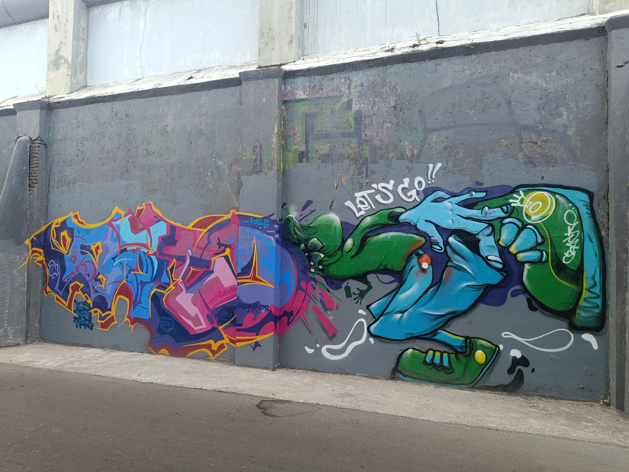The image shows a section of a concrete wall with graffiti art. On the left side of the wall, there is graffiti with vibrant pink, blue, and yellow colors, featuring intricate lettering and abstract shapes. To the right of this, the graffiti depicts two stylized hands, one green and the other blue, with the text "LET'S GO!!" written in white above them. The background of the wall is gray, with some visible wear and tear. Below the wall is a paved area, likely a sidewalk or pathway, and further down is a road surface. The overall setting suggests an urban environment, possibly an underpass or a wall in a public space in the city of Yogyakarta, Indonesia.