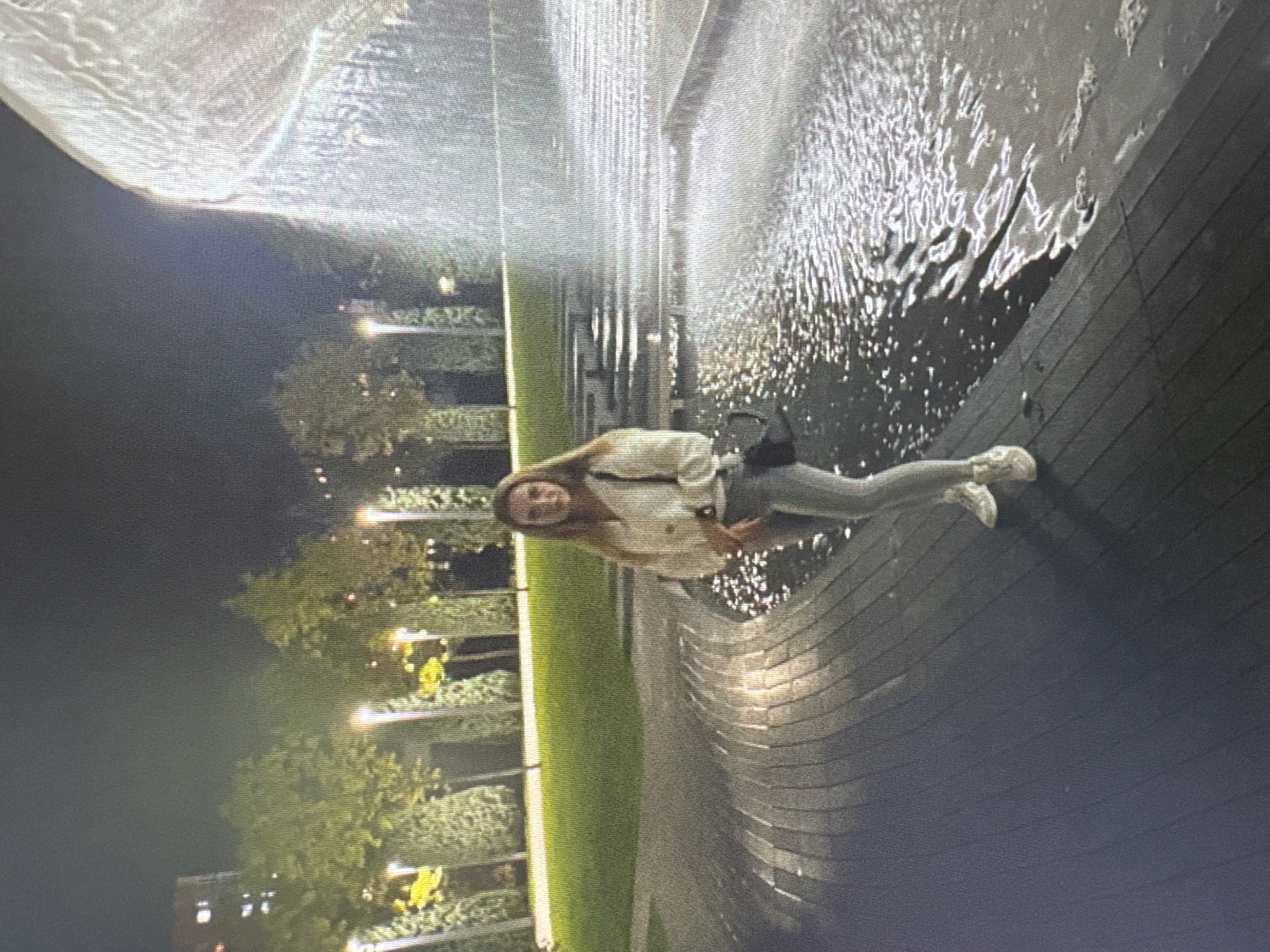 A young woman stands near a fountain in an urban park at night. She is wearing a white cardigan over a light-colored top, gray pants, and white sneakers. She is holding a black purse. The fountain features cascading water illuminated by artificial lights, creating a sparkling effect. In the background, there are trees with lights and a building. The ground around the fountain is made of dark wood with a textured pattern. The scene suggests a public space designed for evening recreation.