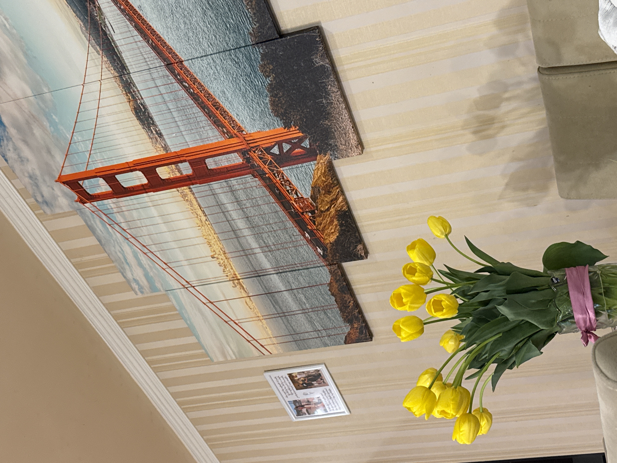 The image shows a room interior with a beige striped wall. On the wall, there is a multi-panel picture of the Golden Gate Bridge in San Francisco. Below the bridge picture, a small framed photograph is present. To the right, a bouquet of yellow tulips in a clear vase with a pink ribbon sits beside a beige couch. The location is unknown and there is no information to suggest Yerevan, Armenia.