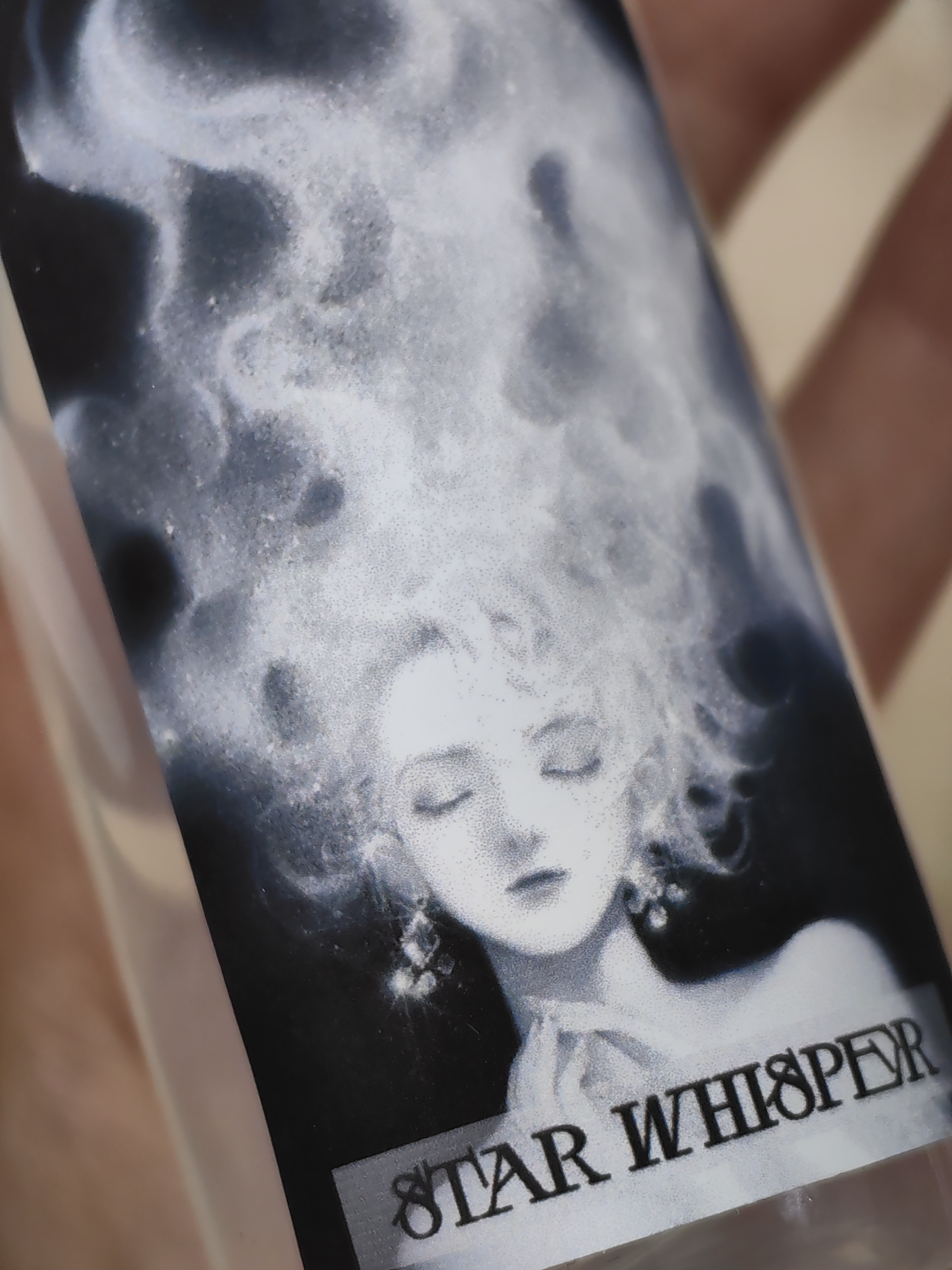 A close-up shot focuses on a portion of a cylindrical object, possibly a bottle or container. A stylized monochrome image is printed on the surface. It depicts a female figure with eyes closed and ethereal, cloud-like hair. She wears delicate earrings and holds her hands clasped together. The text "STAR WHISPER" is printed below the figure. The location is not identifiable from the image.