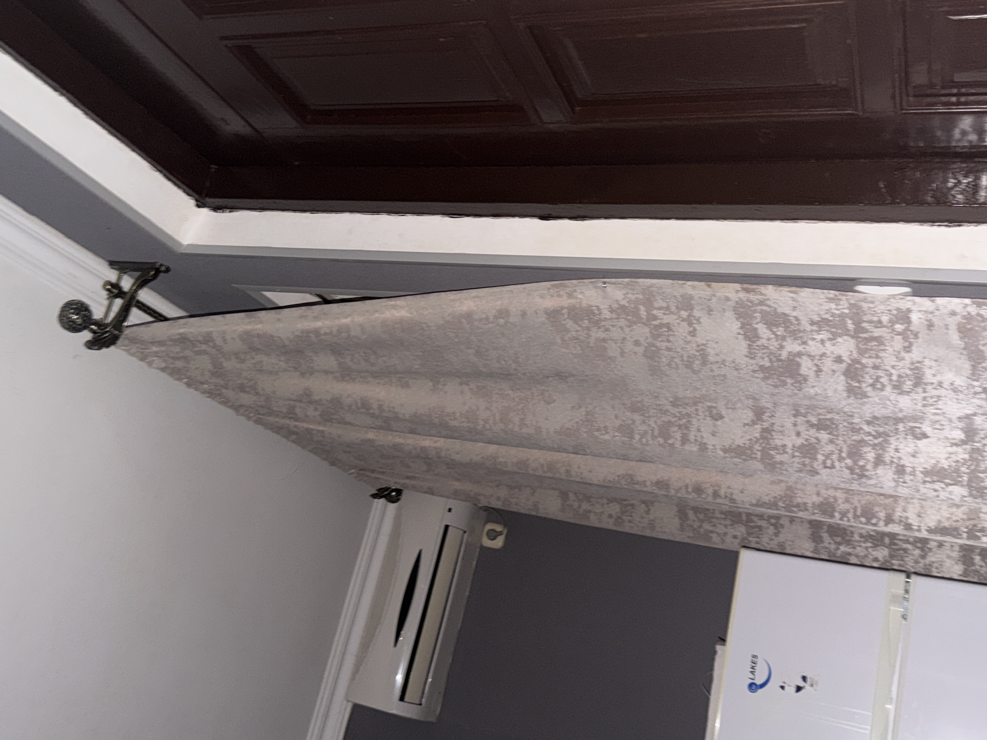 A large, textured, beige curtain hangs from a decorative metal curtain rod attached to a white crown molding. The curtain is draped to the right, partially obscuring a dark brown wooden door. To the left of the curtain, mounted on a grey wall, is a white air conditioning unit. Below the air conditioner, a white refrigerator with a "LAKES" logo is visible. The scene is indoors, with a white ceiling and grey walls. The lighting is dim.