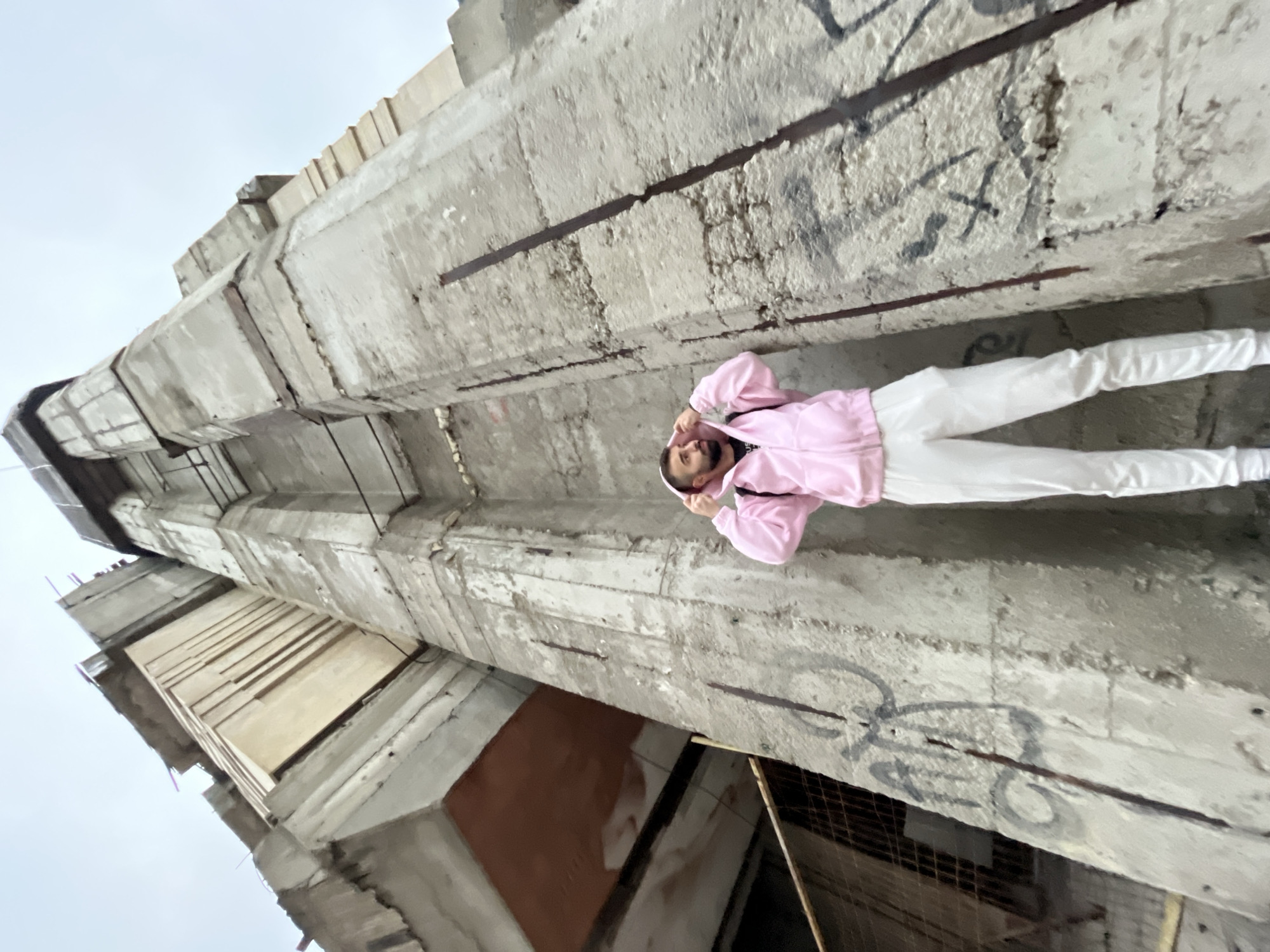 In the city of Unknown, Unknown, a low-angle shot captures a male individual standing before a towering, unadorned concrete structure under an overcast sky. The subject, a male with a dark beard and dark hair, wears a pink hooded zip-up jacket over a dark shirt, white pants, and white shoes. He is positioned facing slightly to his left, looking upwards, with his hands gripping the hood of his jacket. Backpack straps are visible over his shoulders.
The environment consists primarily of a large, monolithic concrete building, exhibiting a raw, unfinished or decaying Brutalist aesthetic. Exposed rebar protrudes from the weathered concrete surfaces, indicating structural elements or damage. Dark graffiti markings are present on the lower sections of the concrete walls. In the lower-left background, a metal mesh fence or barrier is partially visible. The overall scene suggests an urban setting, possibly a construction site or abandoned industrial area.