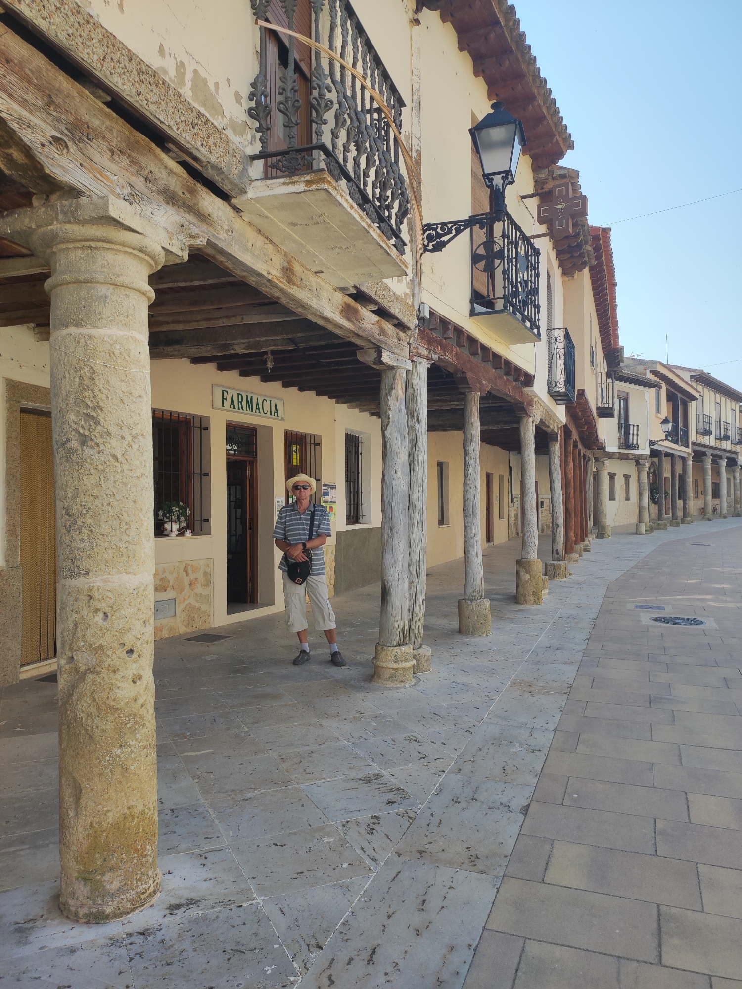 A man wearing a straw hat, striped shirt, and shorts stands in front of a pharmacy with a "FARMACIA" sign. He is holding a bag over his shoulder. The pharmacy is located in a porticoed street with wooden columns and balconies. The street is paved with stone tiles. The scene appears to be in the historic town of Ampudia, Spain, likely on a sunny day.