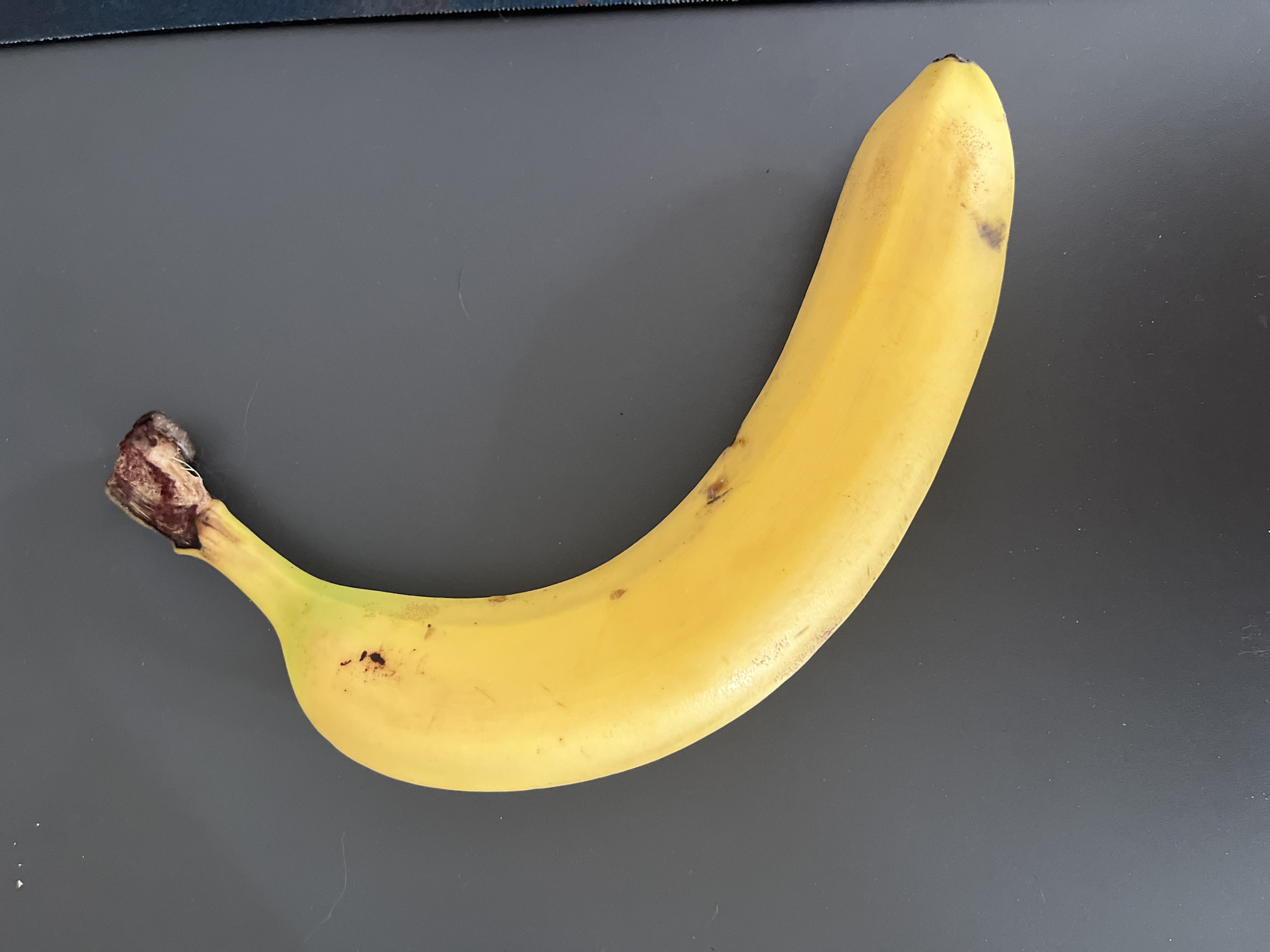 A single, ripe banana is positioned diagonally on a dark, textured surface. The banana is yellow with a few brown spots and a slightly bruised area. The stem end of the banana is darker and appears slightly wilted. The background is a uniform, dark gray color, with subtle dust specks visible upon close inspection. No people or specific landmarks of Shymkent, Kazakhstan are discernible in the image.