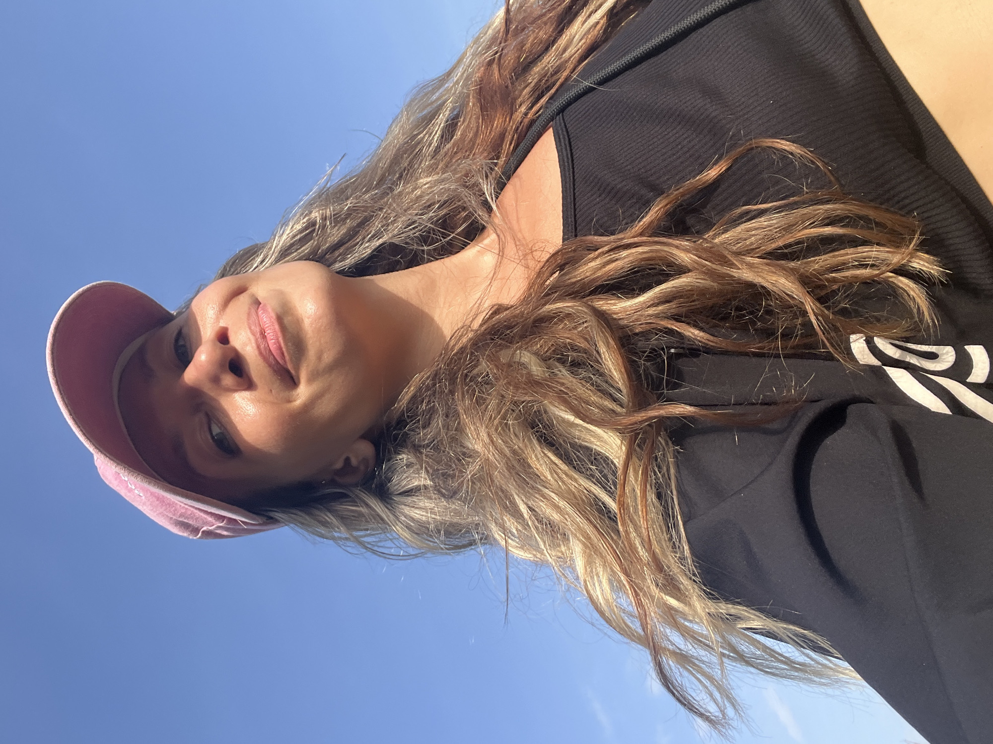 A low-angle, close-up selfie is taken by a woman wearing a pink baseball cap and a black crop top. The woman's face is turned upwards and to the right, with her eyes looking towards the camera. Her hair, which is long and wavy with highlights, is blowing in the wind. The background is a clear blue sky. The image appears to have been taken outdoors, possibly in the city of Talalla, Sri Lanka, given the bright sunlight and clear sky.