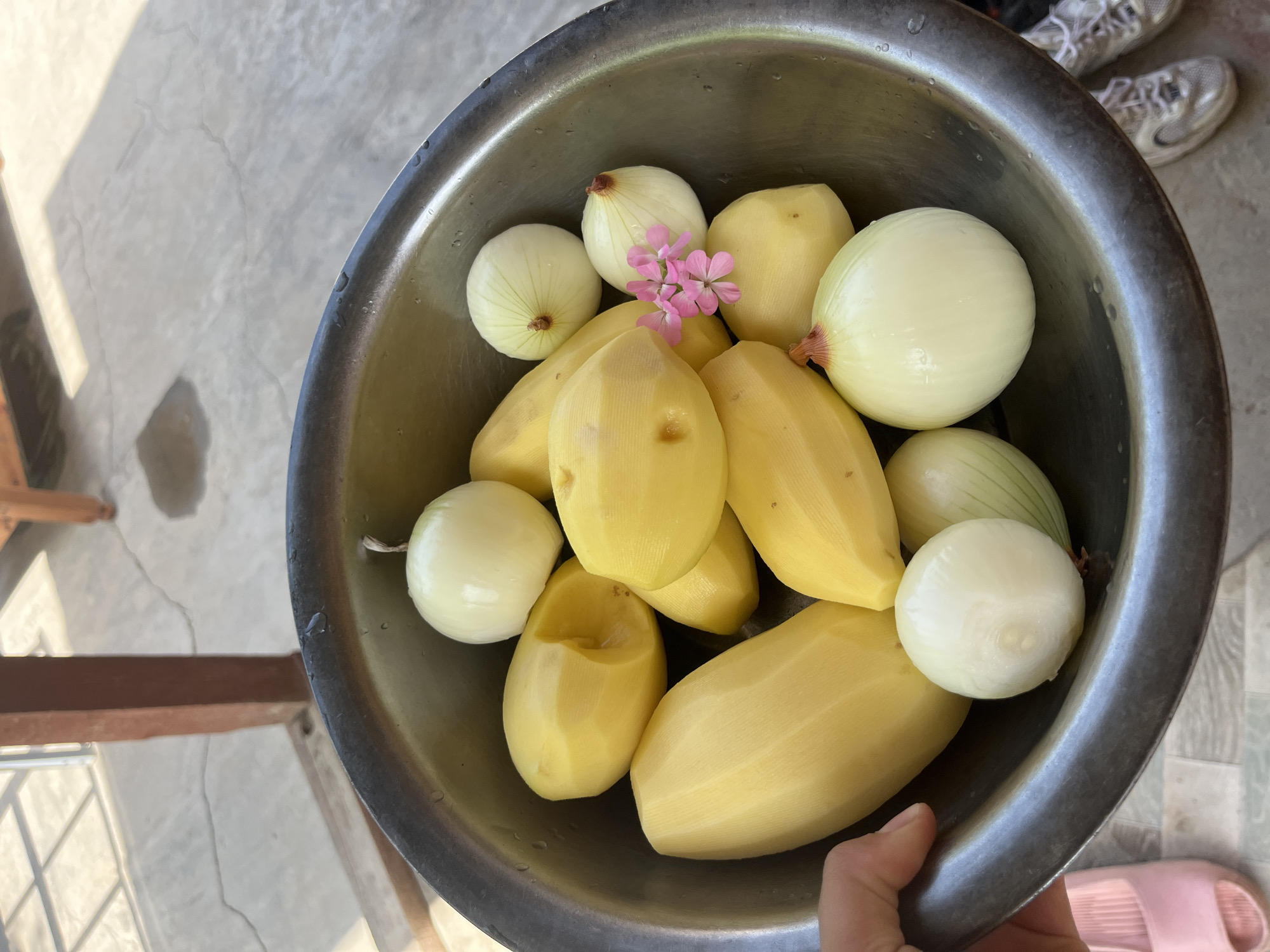 A medium shot captures a metal bowl filled with peeled potatoes and whole onions, with a few delicate pink flowers resting on top of one onion. The bowl is placed outdoors on a concrete surface, with sunlight casting shadows from a wooden structure in the background, suggesting a patio or courtyard setting. The scene is indicative of food preparation, possibly in a residential area in Batken City, Kyrgyzstan. The focus is on the fresh produce, with the surrounding environment subtly hinting at a warm, sunny day.