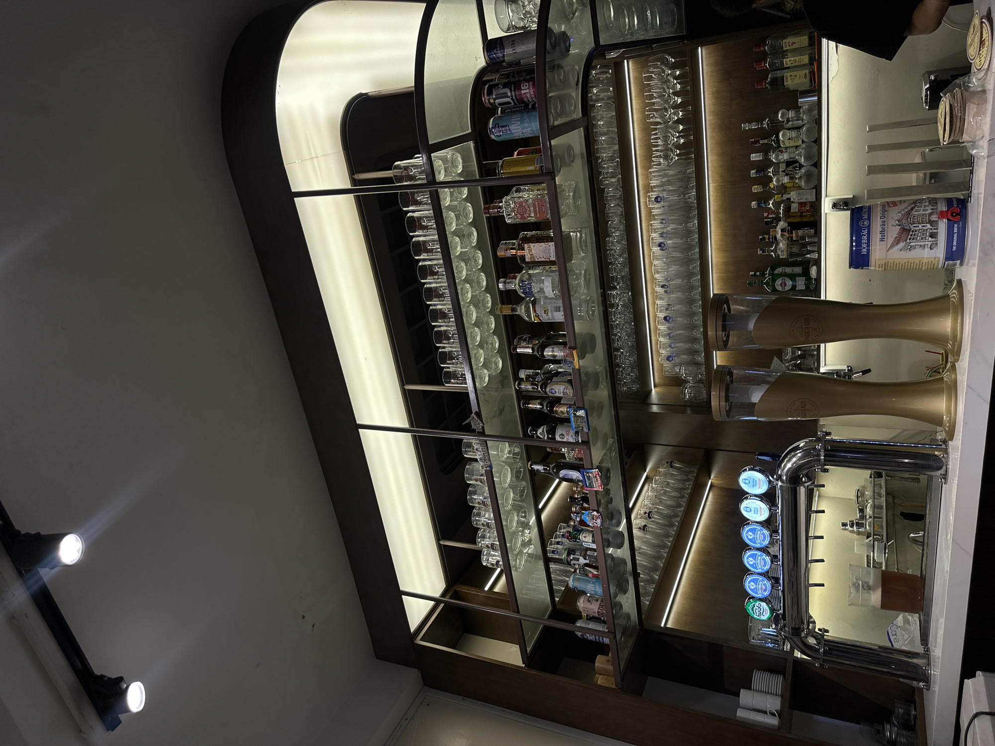 This image presents the interior of a well-appointed bar or pub, likely situated in Nha Trang Ward, Vietnam, given the provided context. The scene is dominated by a meticulously arranged shelving unit showcasing a diverse selection of alcoholic beverages and glassware, bathed in warm, artificial light.
The central feature is a series of multi-tiered shelves constructed from dark wood and reflective panels, possibly glass or polished metal. These shelves are illuminated by integrated strip lighting, highlighting an array of spirit bottles, including Absolut Vodka, gin (with a discernible Bombay Sapphire label), and various whiskies and liqueurs. Alongside these, numerous clean glass items, such as tumblers, pint glasses, and wine glasses, are neatly stacked, ready for service.
On the right, a modern bar counter is equipped for dispensing drinks. Two prominent golden beer taps, partially branded "ARKHAUS," stand tall, accompanied by a sleek chrome tap system featuring multiple spouts. One spout is clearly marked "GUINNESS" with a green illuminated emblem, while others display blue lights, suggesting different beer options. A cylindrical beverage cooler on the counter prominently displays "Hofbräu München Original Weiss," indicating a European beer offering. Stacked coasters and a plastic container labeled "CORE" are also visible on the counter.
Overhead, bright white spotlights on a track provide additional illumination, complementing the softer, ambient glow from curved, recessed light fixtures positioned along the wall above the shelving. The setting suggests an evening or nighttime ambiance, as indicated by the reliance on artificial lighting. No individuals are visible, but the pristine setup implies readiness for customer service. The overall aesthetic is modern, clean, and inviting.