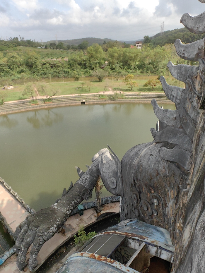 The image captures an elevated perspective of an outdoor scene in Thủy Xuân Ward, Vietnam. In the immediate foreground, to the right, is a large, weathered sculptural structure, predominantly gray with visible patches of rust and faded blue paint. This structure features a prominent, scaled appendage resembling a creature's hand or claw extending downwards towards the water. Below this, a section of a dilapidated platform or roof with exposed framing and two small, dark solar panels is visible, along with graffiti on its surfaces. The main body of the structure displays textured, layered forms and circular relief patterns.
Below the foreground structure, a large, still body of greenish-brown water occupies the midground. Its edge is lined with multiple tiers of concrete retaining walls, where a dark, rectangular culvert opening is present. Beyond the water, a strip of green grass is visible, interspersed with various trees, some with sparse foliage, and dirt paths. The background features rolling hills covered in dense green vegetation. A distant, light-colored, multi-story building and a metal power transmission tower are discernible on these hills. The sky is overcast with scattered clouds. No individuals are present in the scene.